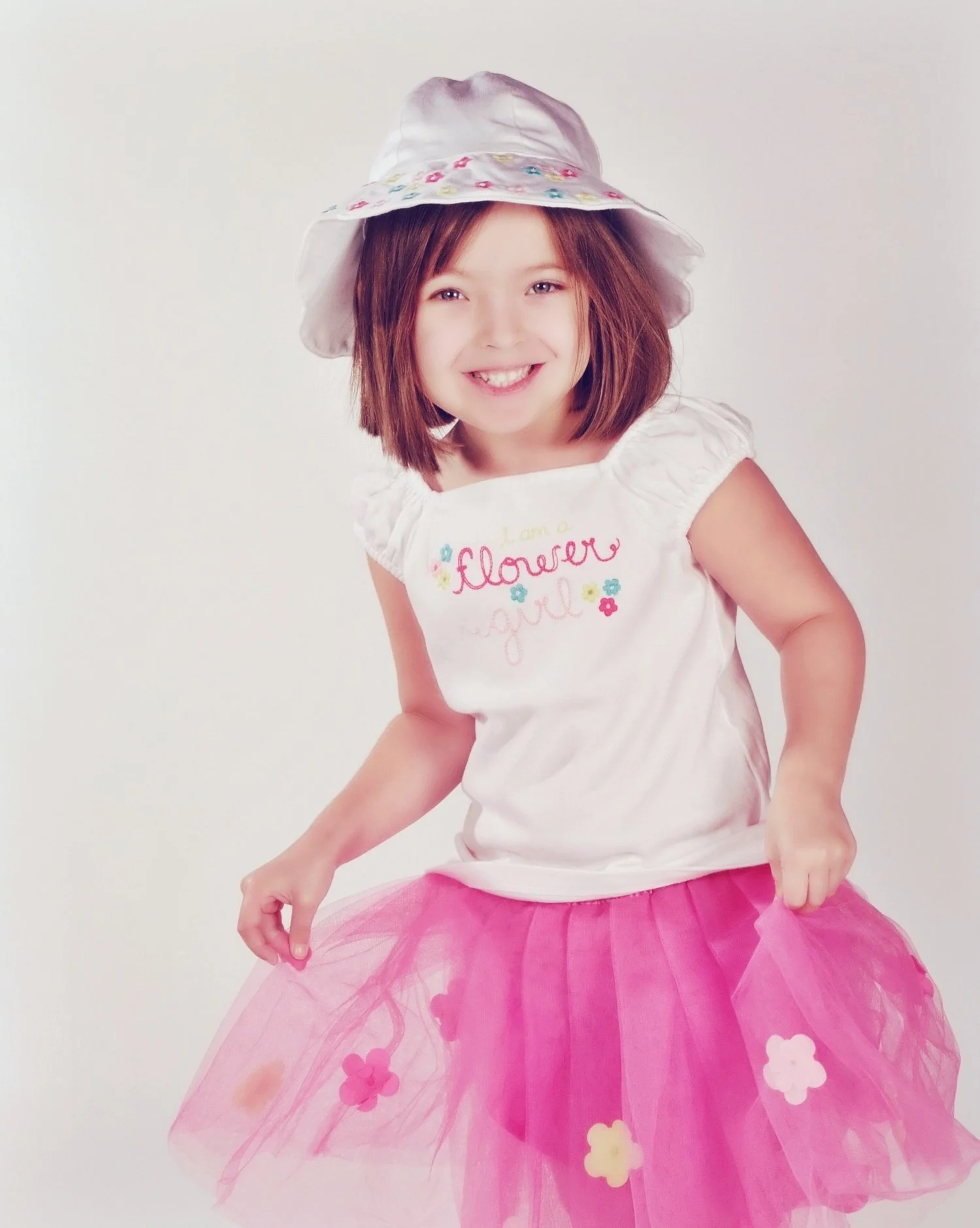 A young girl with shoulder-length brown hair is smiling, wearing a white sunhat with colorful embroidery, a white shirt with floral embroidery that says 'I am a flower,' and a pink tulle skirt with flower appliqués, standing against a plain white bac