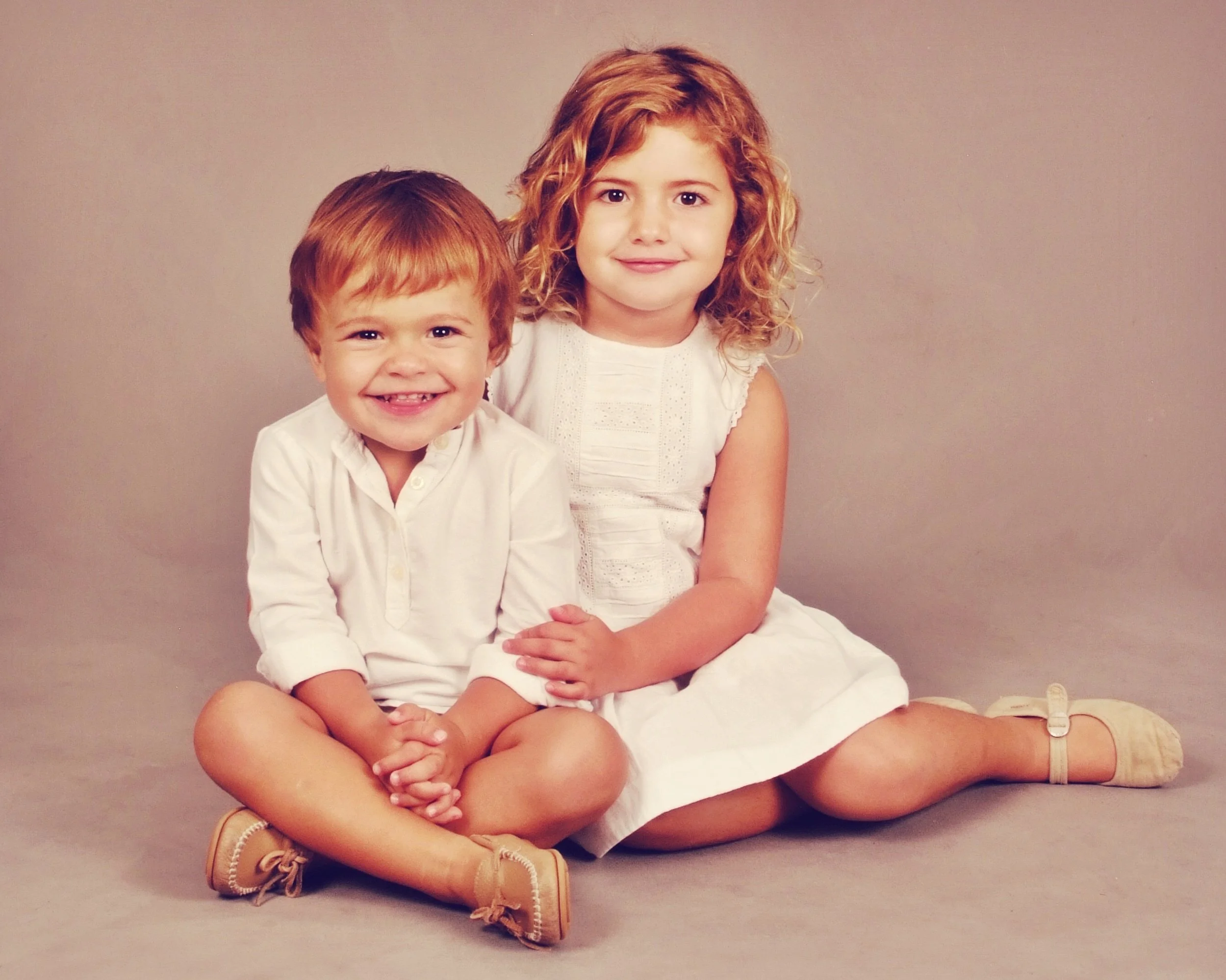 Two young children, a boy and a girl, sitting on the floor against a plain beige background. The girl has curly red hair and is smiling softly, and the boy has straight red hair and is showing a big smile with missing front teeth. Both are wearing wh