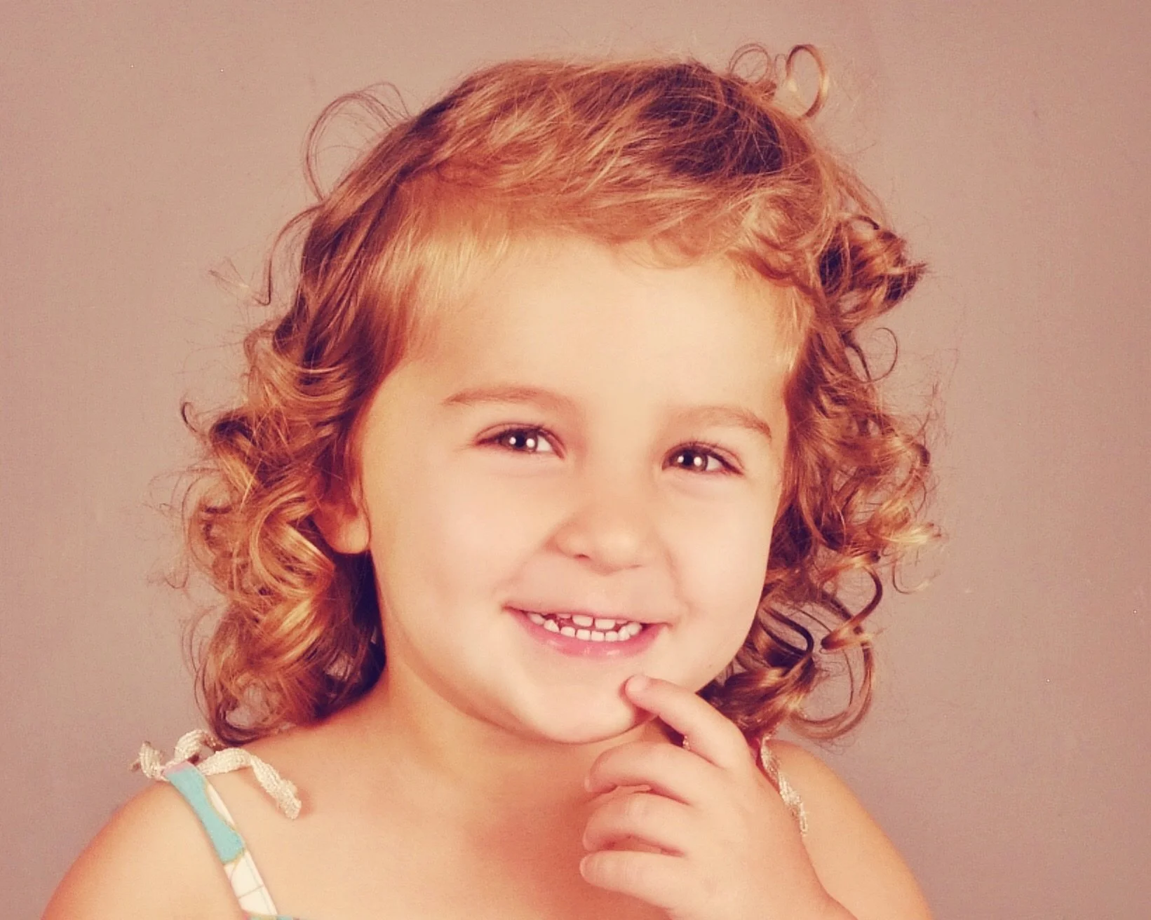 A young girl with curly red hair, smiling and touching her chin with her finger, wearing a light blue dress with white ribbon straps.