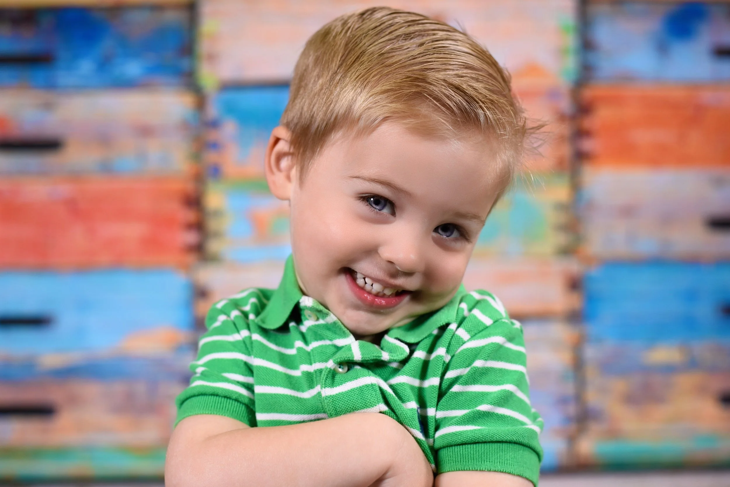 Close-up of a young boy with blonde hair and blue eyes, smiling and crossing his arms, wearing a green and white striped shirt, with a blurry colorful background.