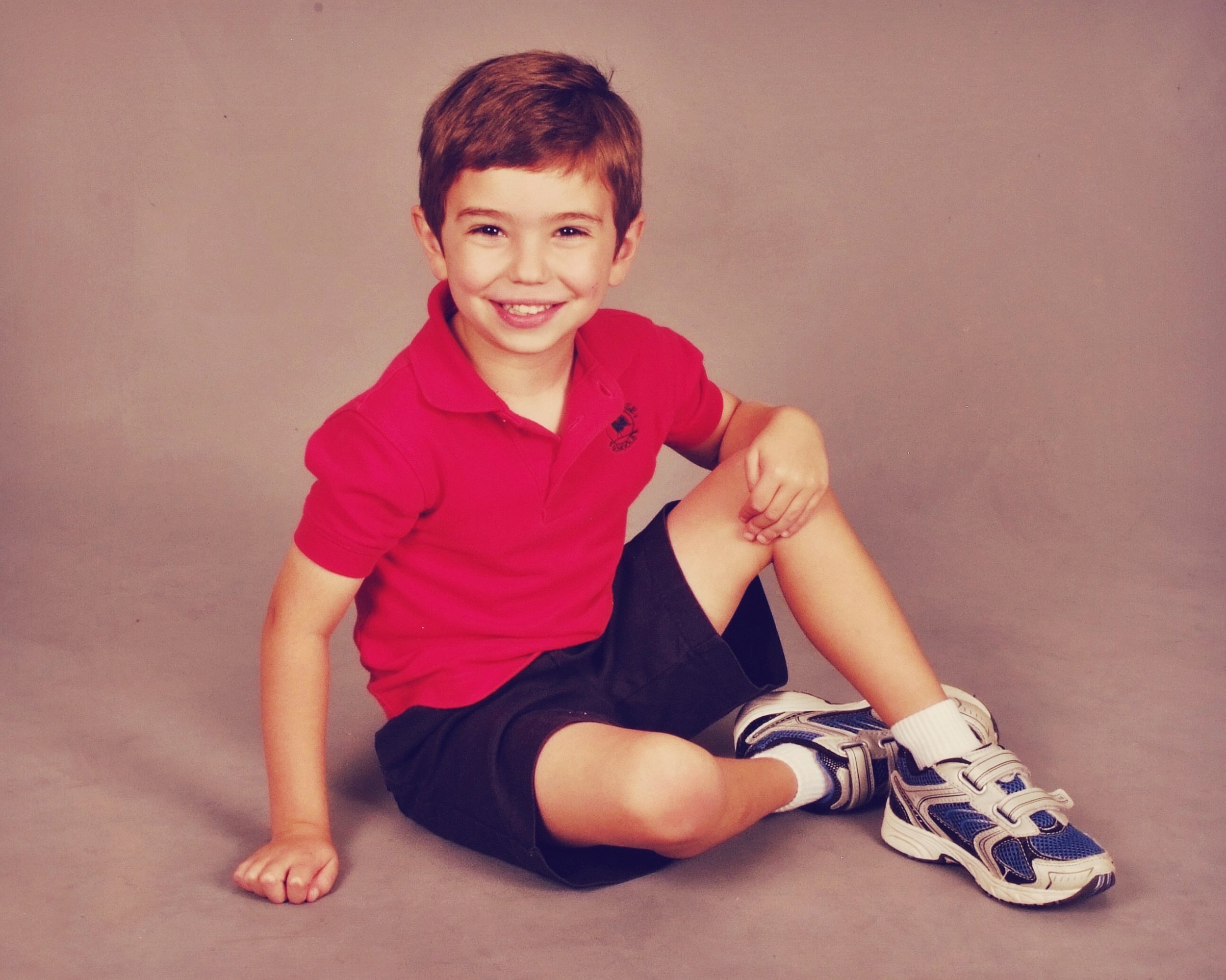 A young boy sitting on the floor, smiling, wearing a red polo shirt, dark shorts, white socks, and athletic shoes against a plain background.