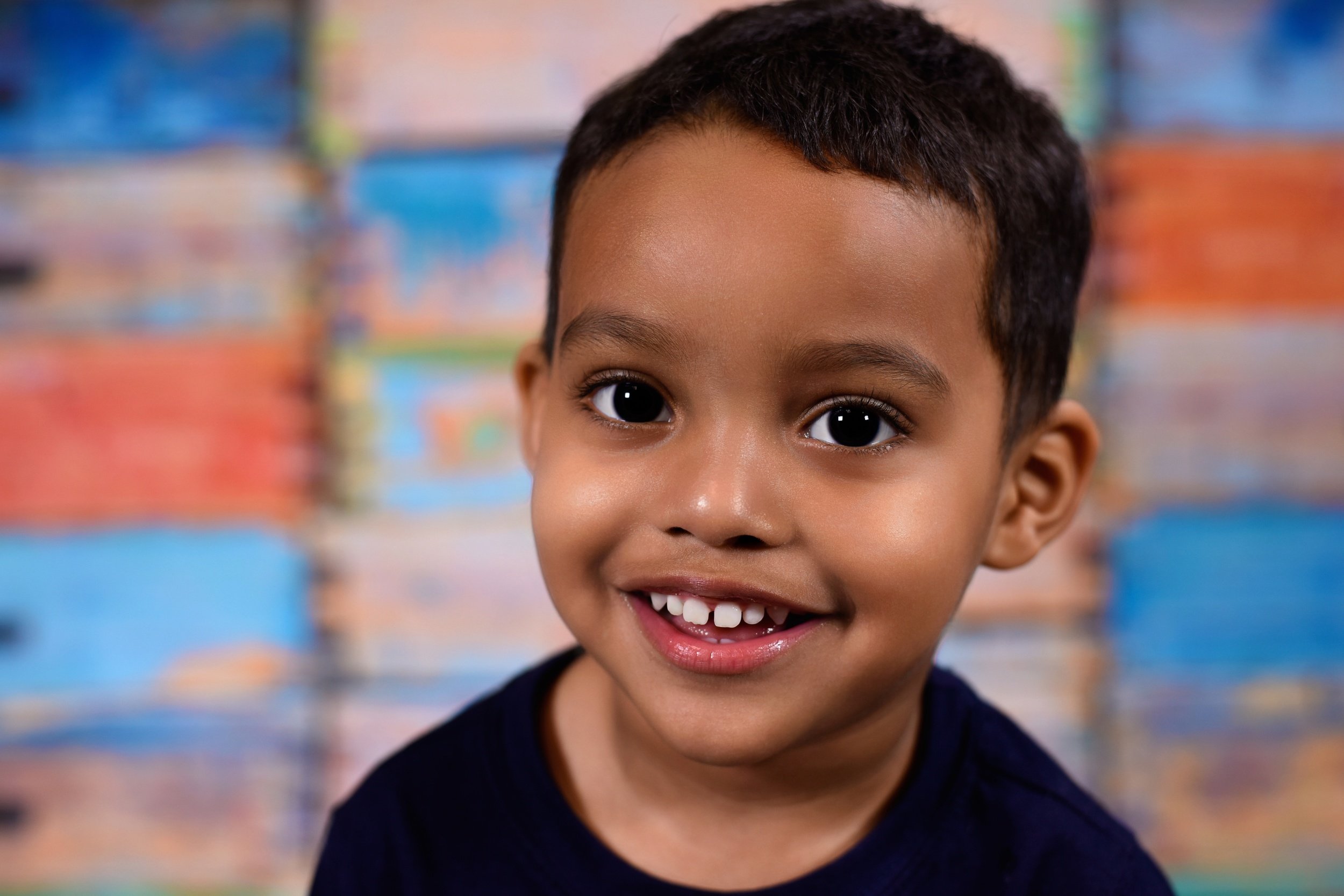 A young boy with short dark hair, big dark eyes, and a wide smile showing his teeth, wearing a dark shirt, in front of a colorful, out-of-focus background.