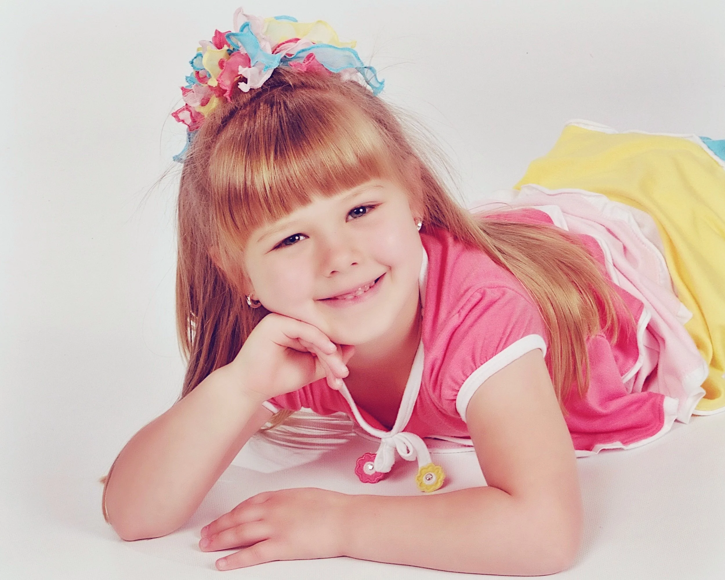 A young girl with long red hair, wearing a pink top with white and pink details, lying on her side with her head resting on her hand, smiling at the camera.