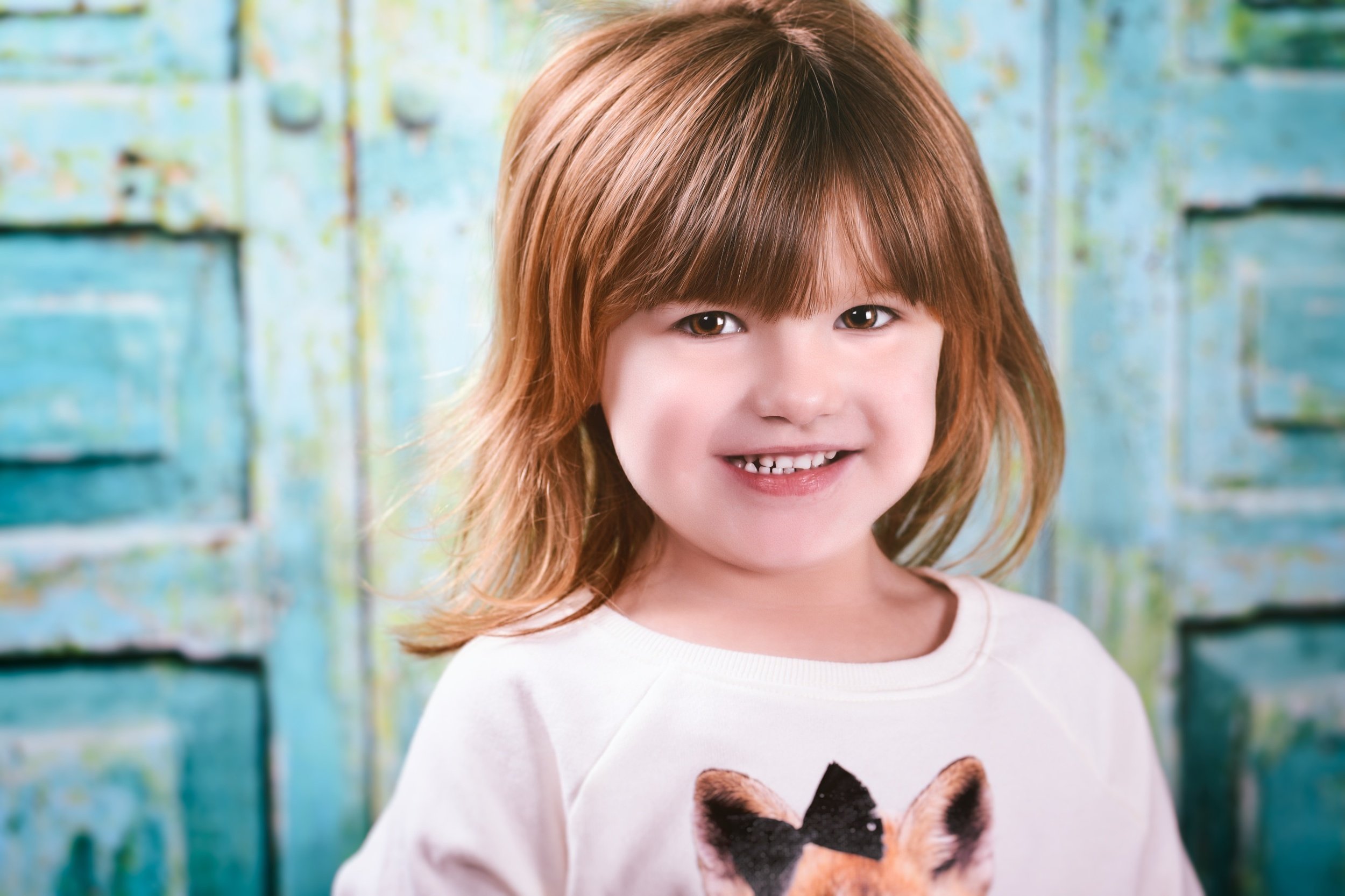 A young girl with red hair smiling, wearing a white shirt with a puppy graphic on it, standing outdoors in front of a blue painted wooden fence.