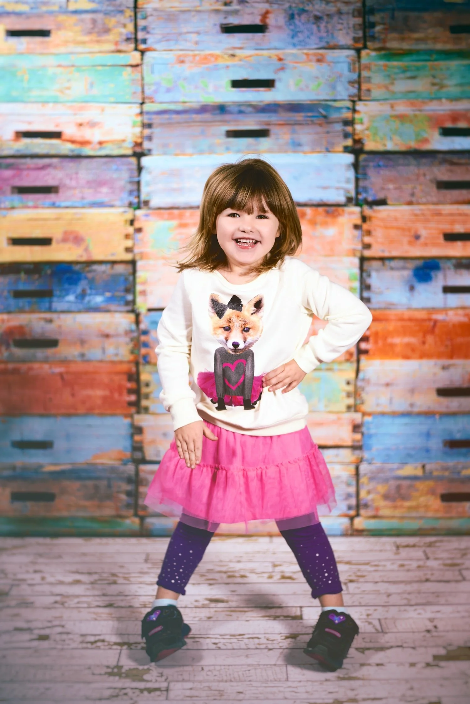 Smiling young girl with brown hair, wearing a cream-colored sweater with a fox graphic, a pink skirt, purple tights with white polka dots, and sneakers, standing with legs apart and hand on hip in front of a colorful wooden wall.