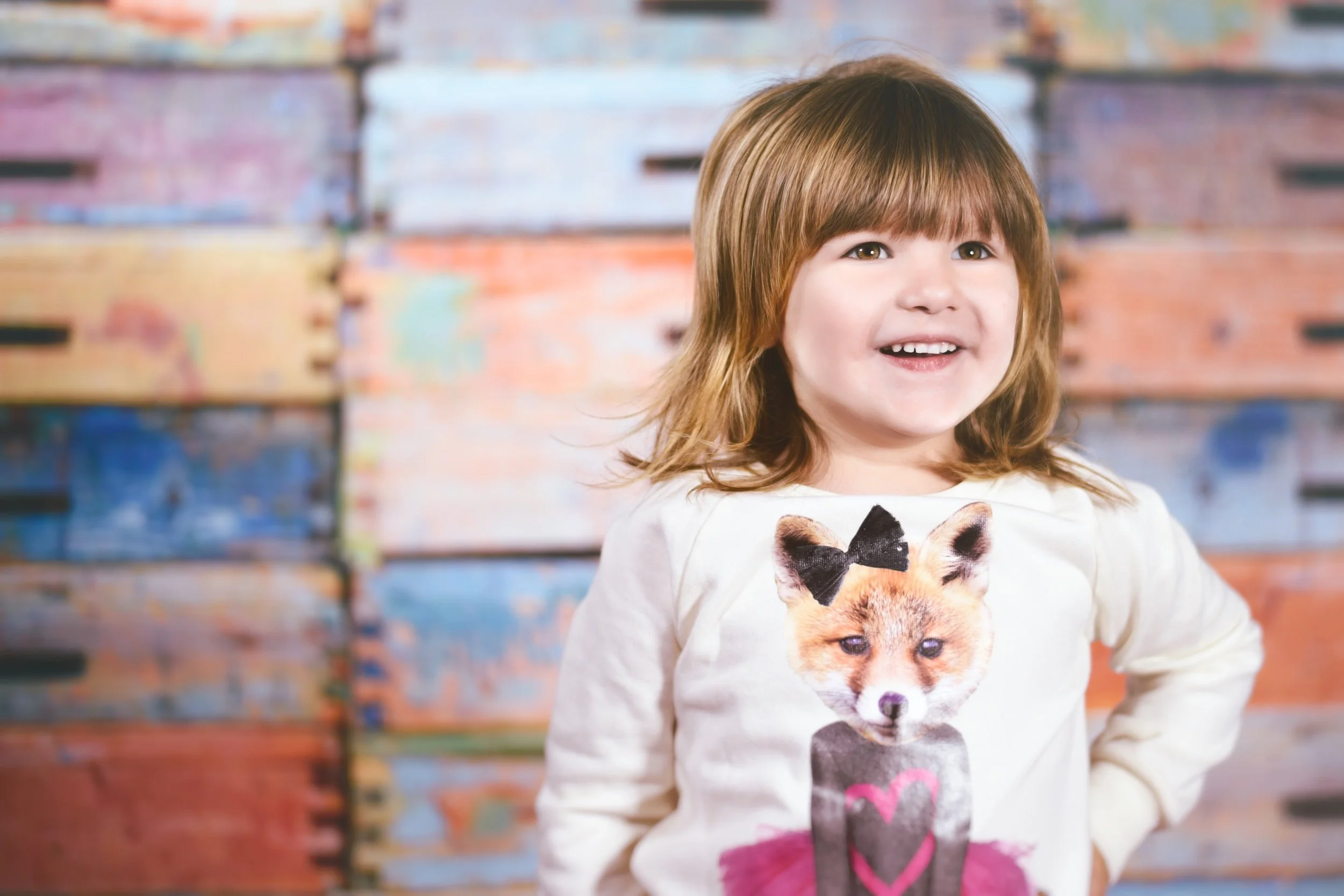 A young girl with brown hair and brown eyes smiling, wearing a white sweatshirt with a puppy face print, standing in front of a colorful, abstract painted wall.