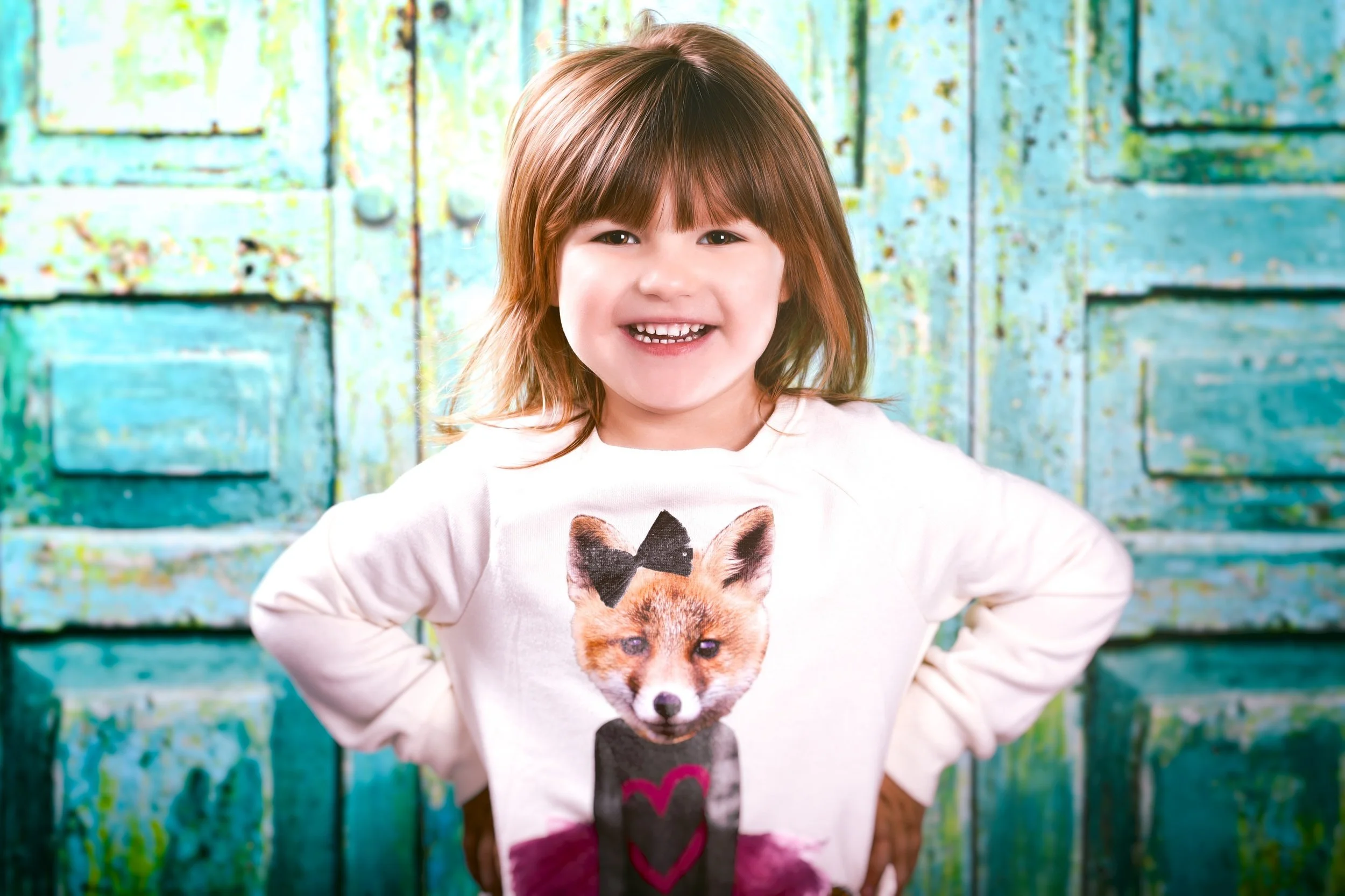 A young girl with brown hair smiling and wearing a white shirt with a dog graphic, standing in front of a blue, weathered wooden background.