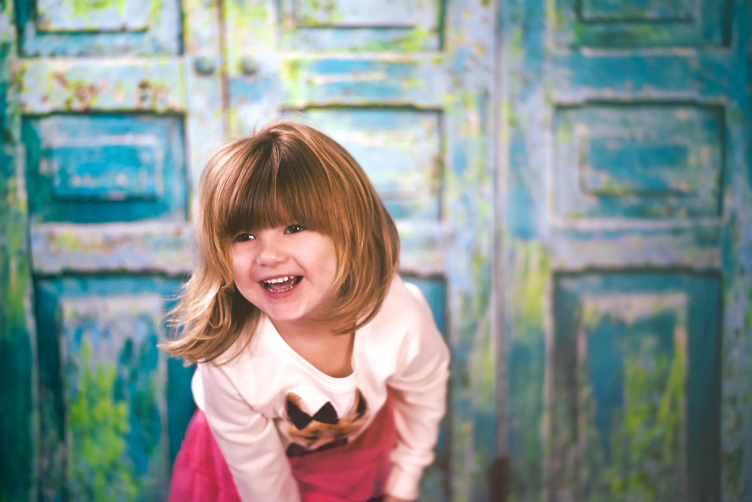 A young girl with red hair smiling and leaning forward, in front of a colorful, abstract painted background.