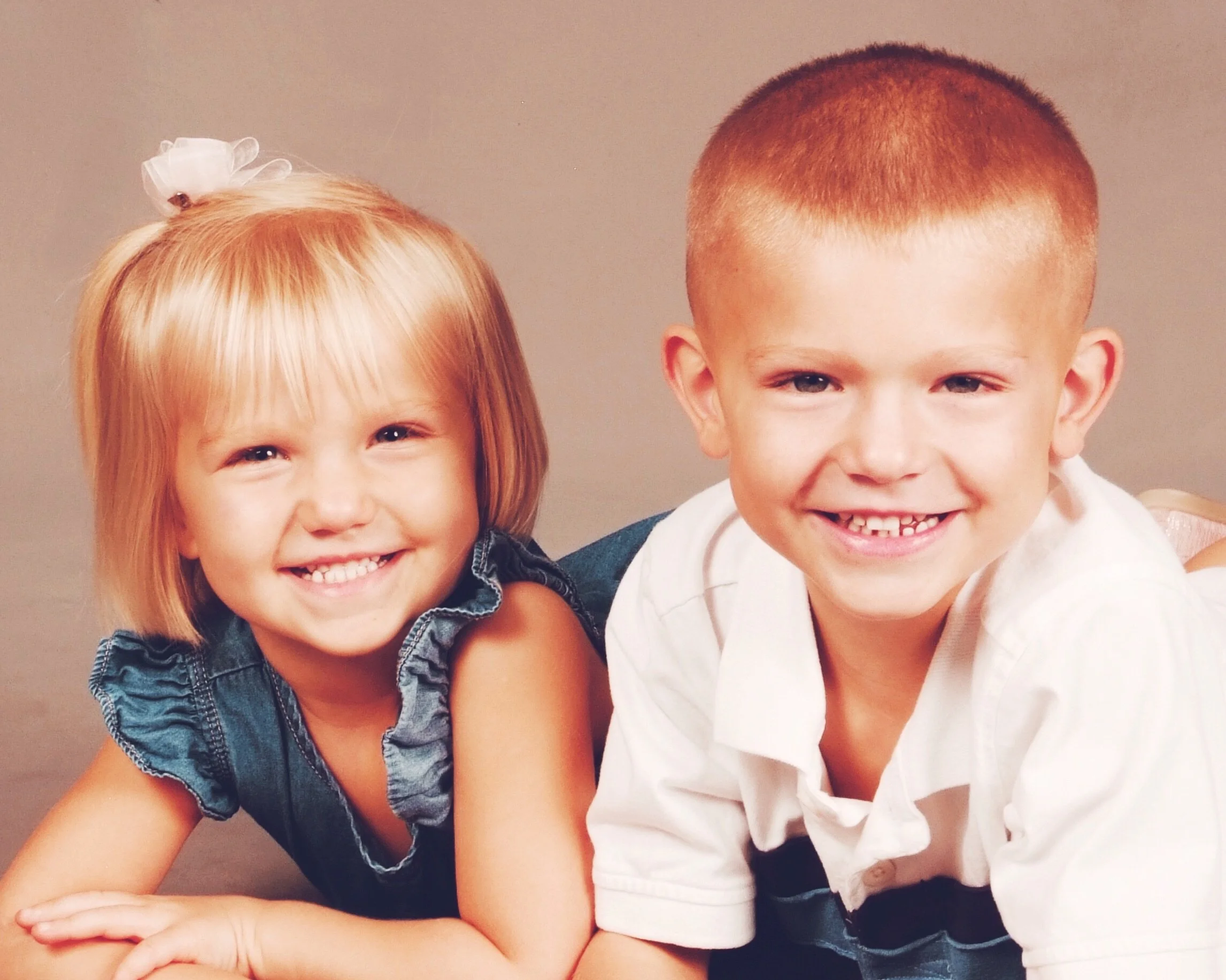 Two young children, a girl and a boy, smiling and close together in a portrait. The girl has blonde hair with a big white bow and is wearing a denim top. The boy has a crew cut hairstyle and is wearing a white shirt.