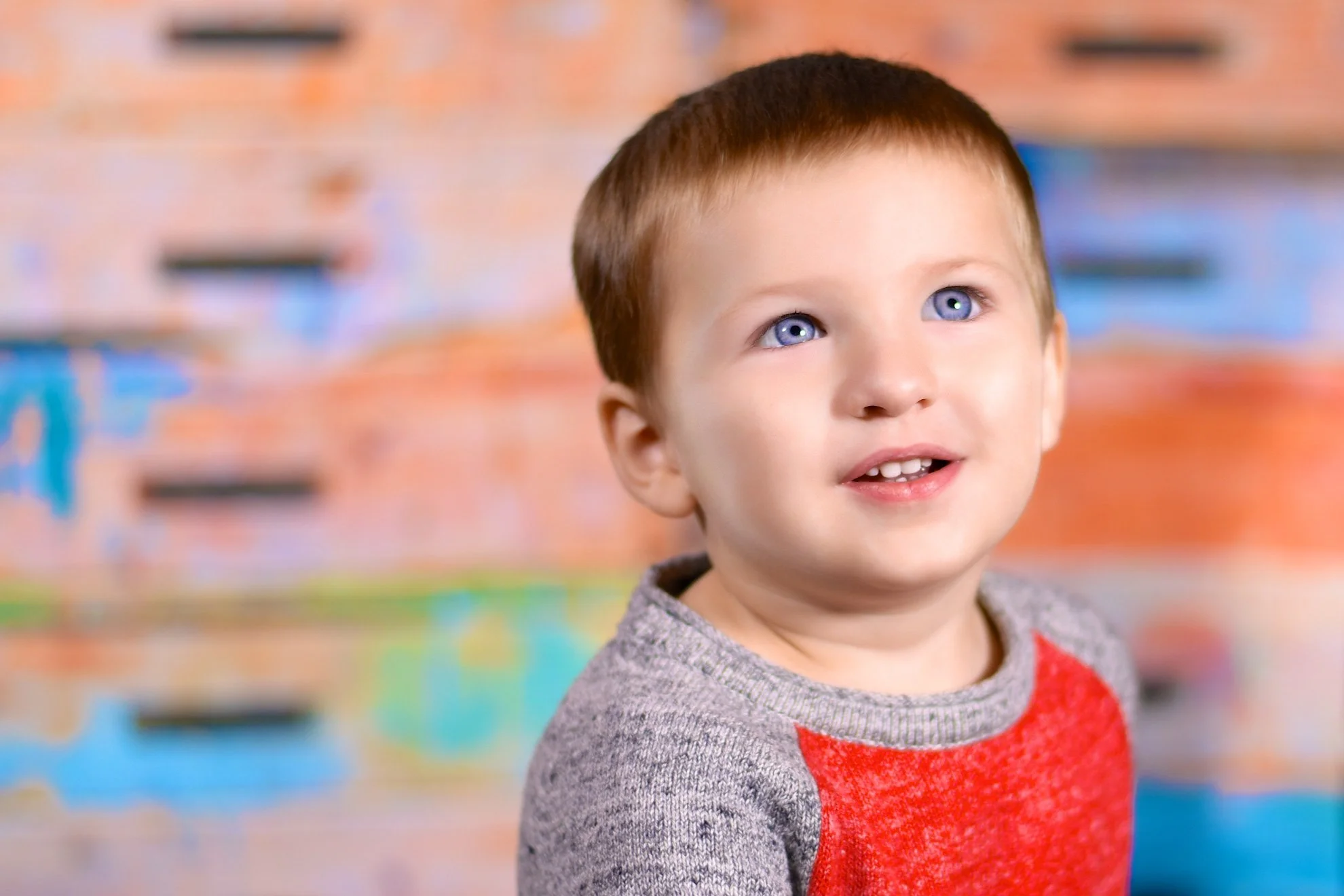 Young boy with short brown hair and blue eyes looking upwards, wearing a gray and red sweater, standing in front of a colorful, abstract background.