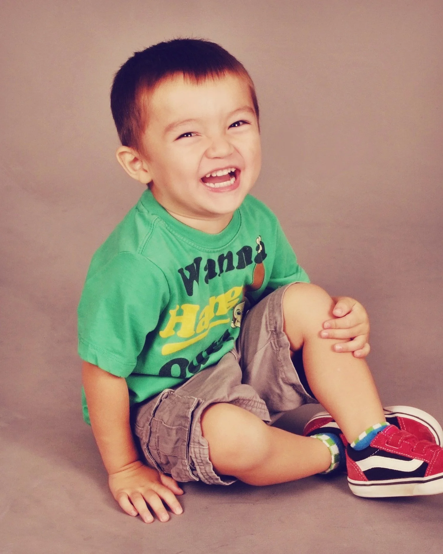 A young boy with short hair, wearing a green T-shirt with yellow and black text, khaki shorts, and red sneakers, sitting on the floor and laughing with his mouth open, showing his teeth, against a plain gray background.