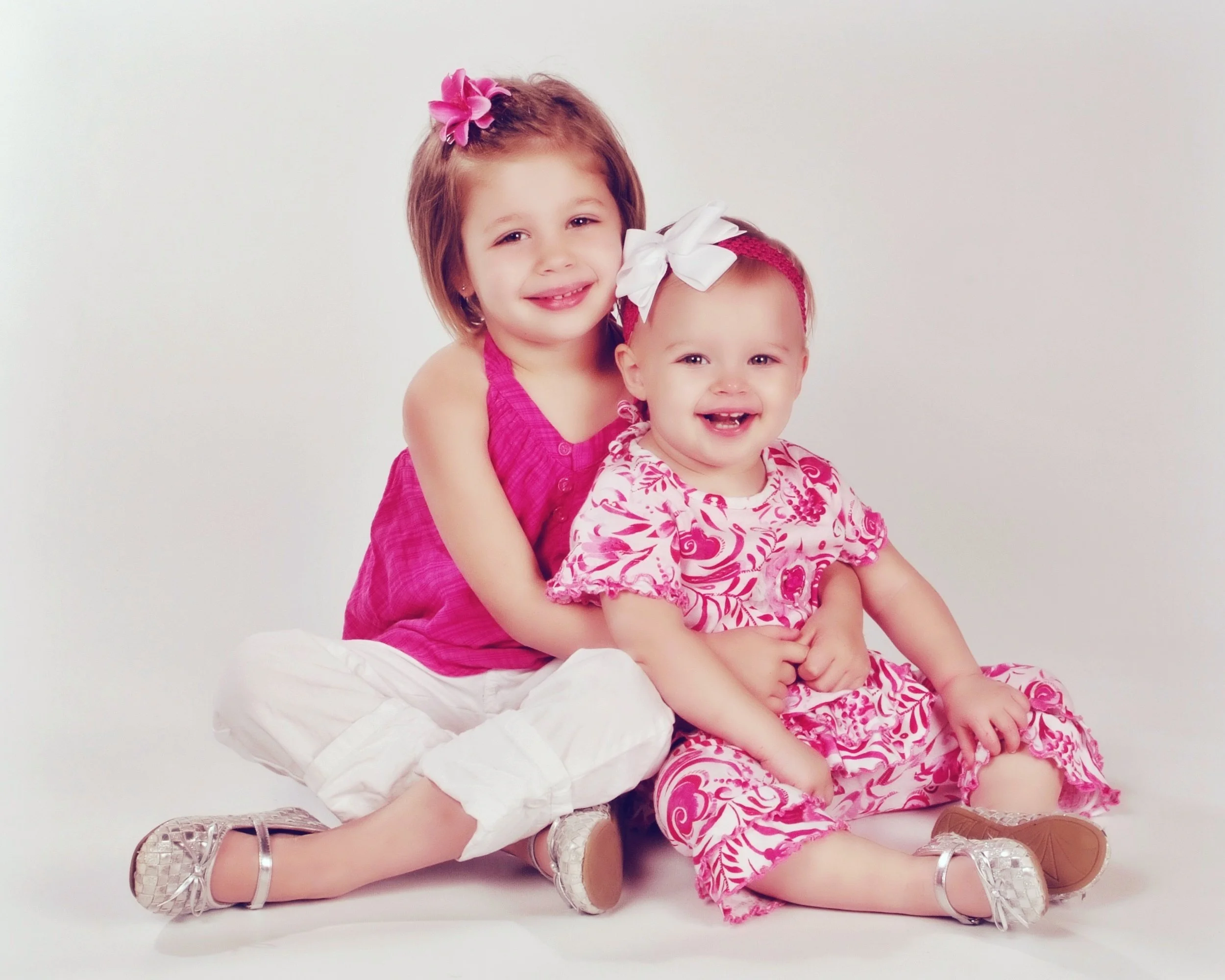 Two young girls sitting together, smiling. The older girl has brown hair with a pink flower hair clip, and the younger girl has a white bow in her hair. Both are wearing pink patterned dresses. The older girl has her arms around the younger girl, who