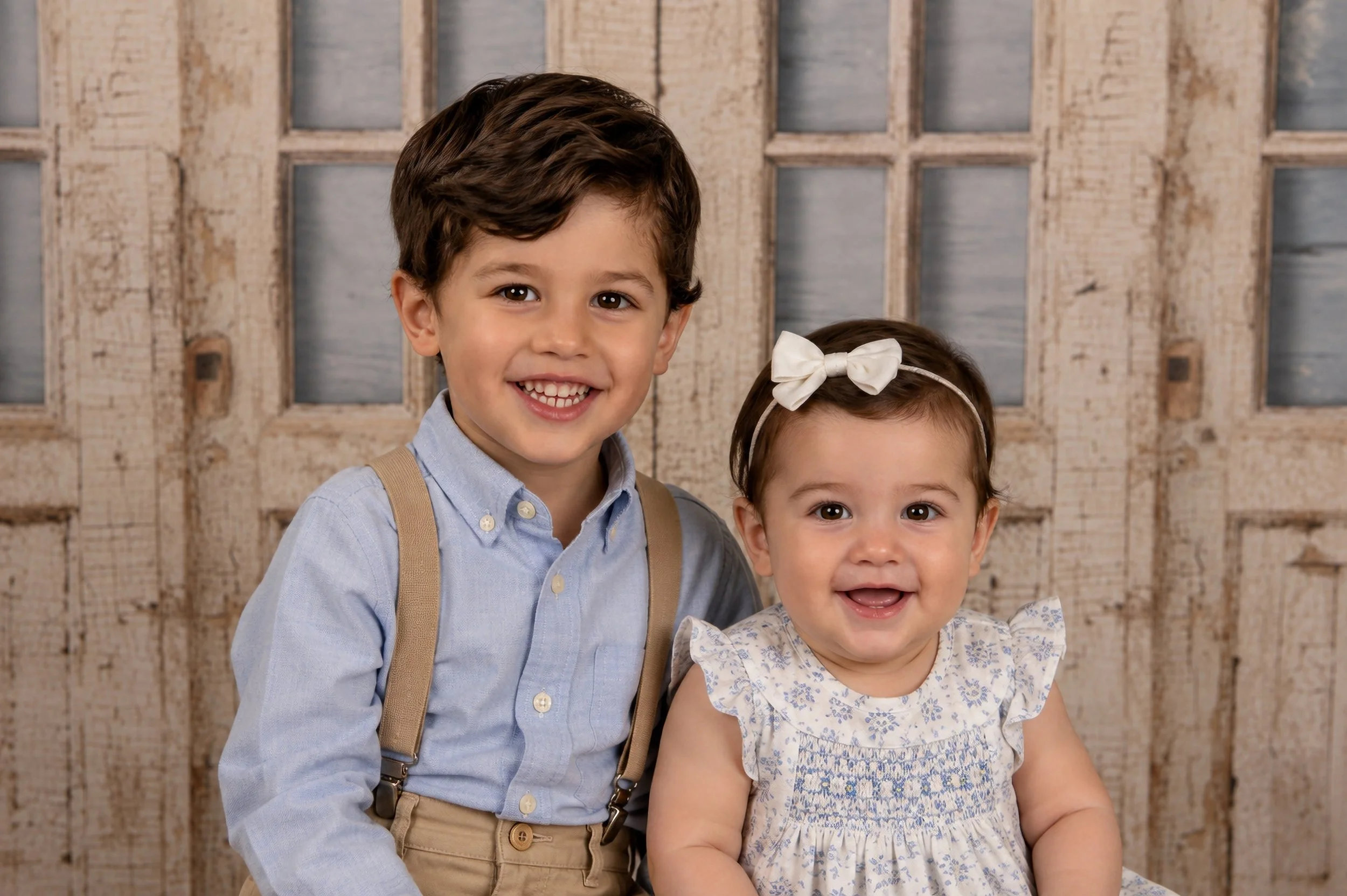 Two young children smiling in front of a rustic wooden backdrop. The boy has brown hair, wears a light blue shirt with suspenders, and the girl has brown hair with a white bow, dressed in a floral patterned shirt.