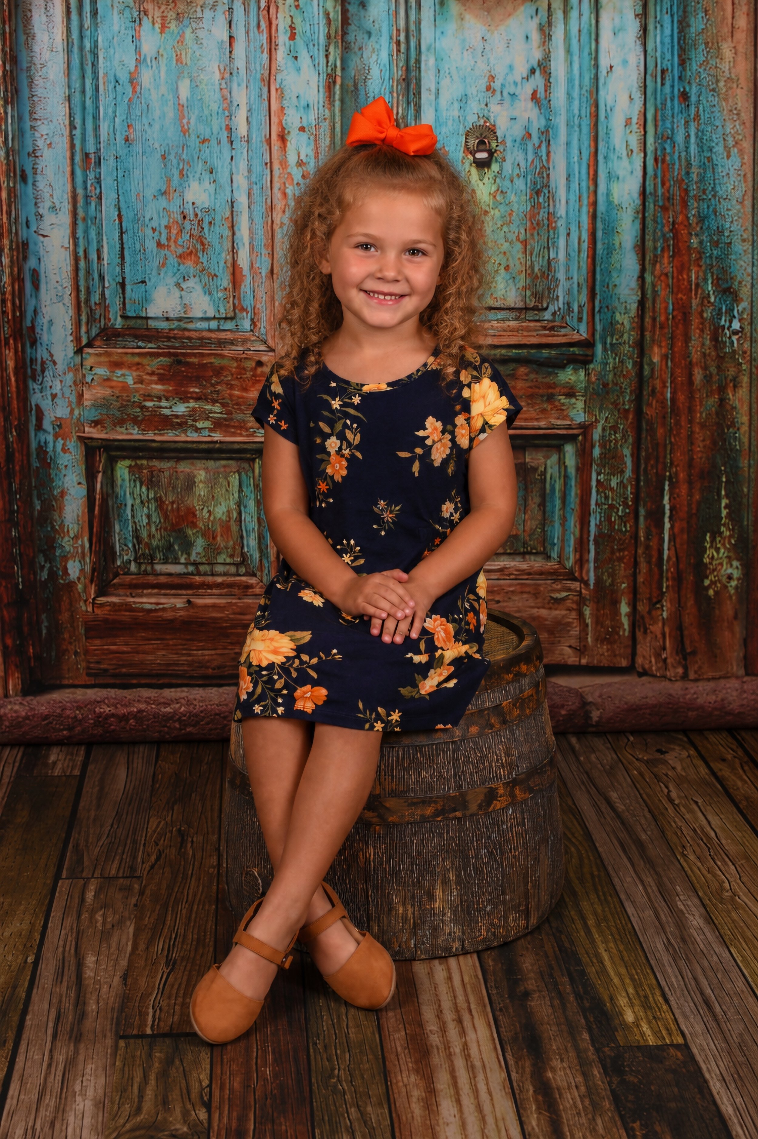 Young girl sitting on a barrel, smiling, wearing a navy floral dress with an orange bow in her curly hair, in front of a rustic blue and brown wooden door background.