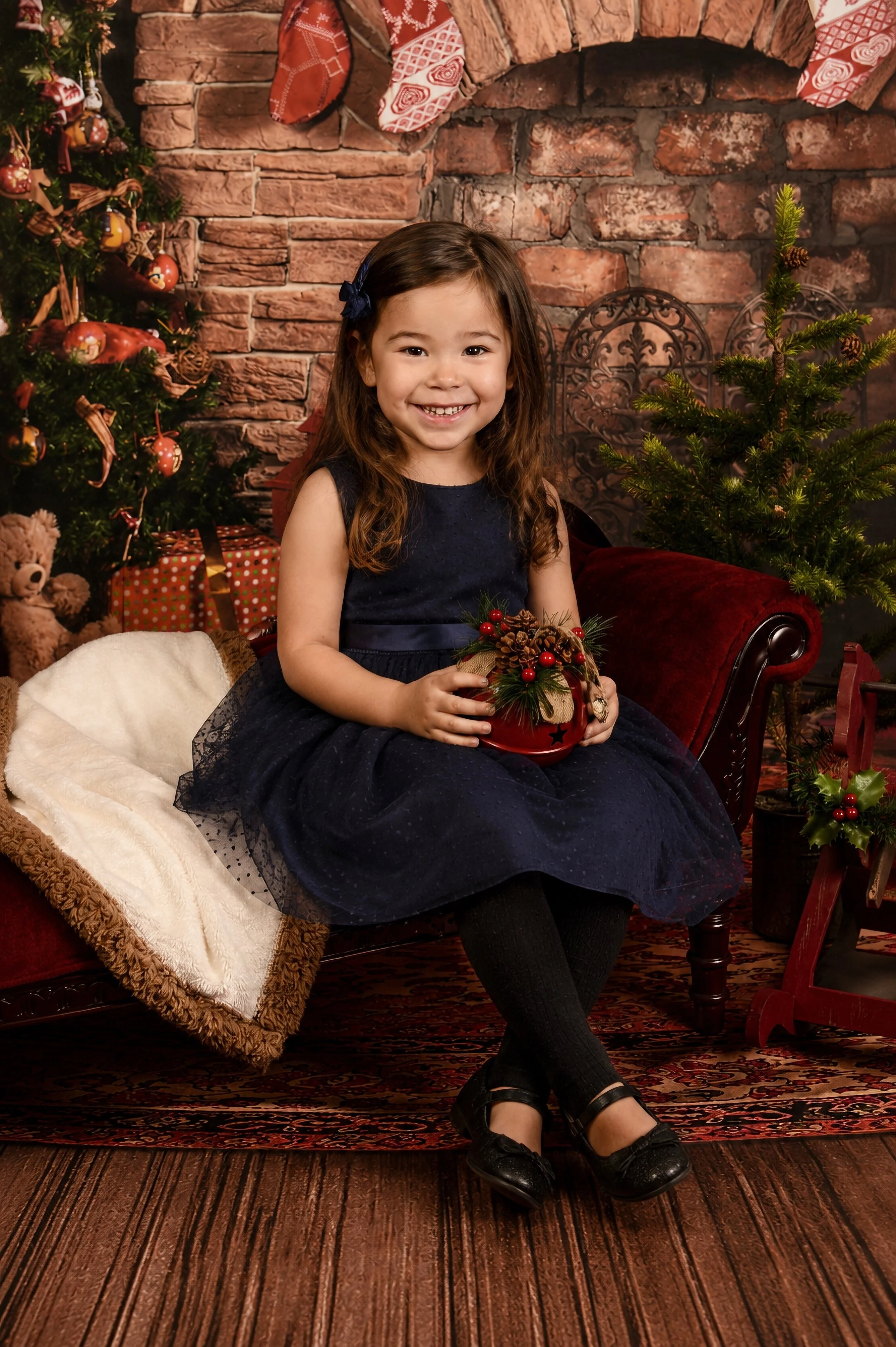 A young girl sitting on a vintage wooden chair in front of a brick fireplace decorated with Christmas stockings hanging from the mantle. She is smiling, holding a holiday-themed centerpiece, and wearing a navy blue dress with black shoes. To her righ