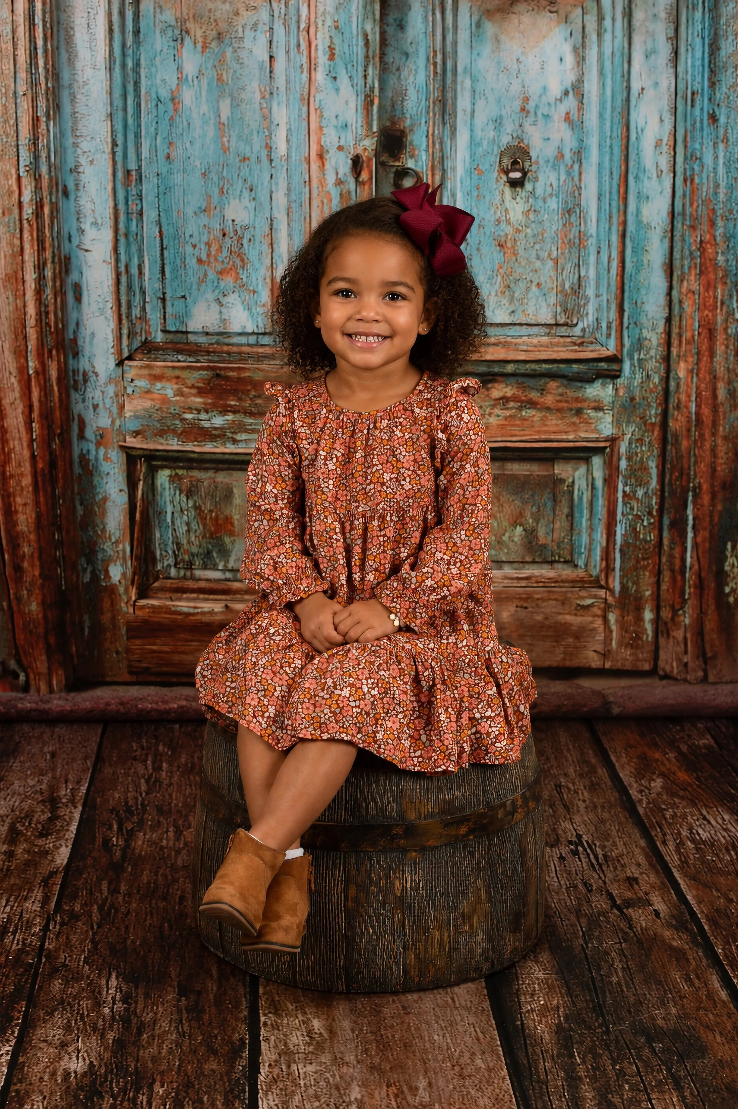 A young girl with curly hair, wearing a floral dress and brown boots, smiling and sitting on a wooden barrel in front of a rustic wooden background.