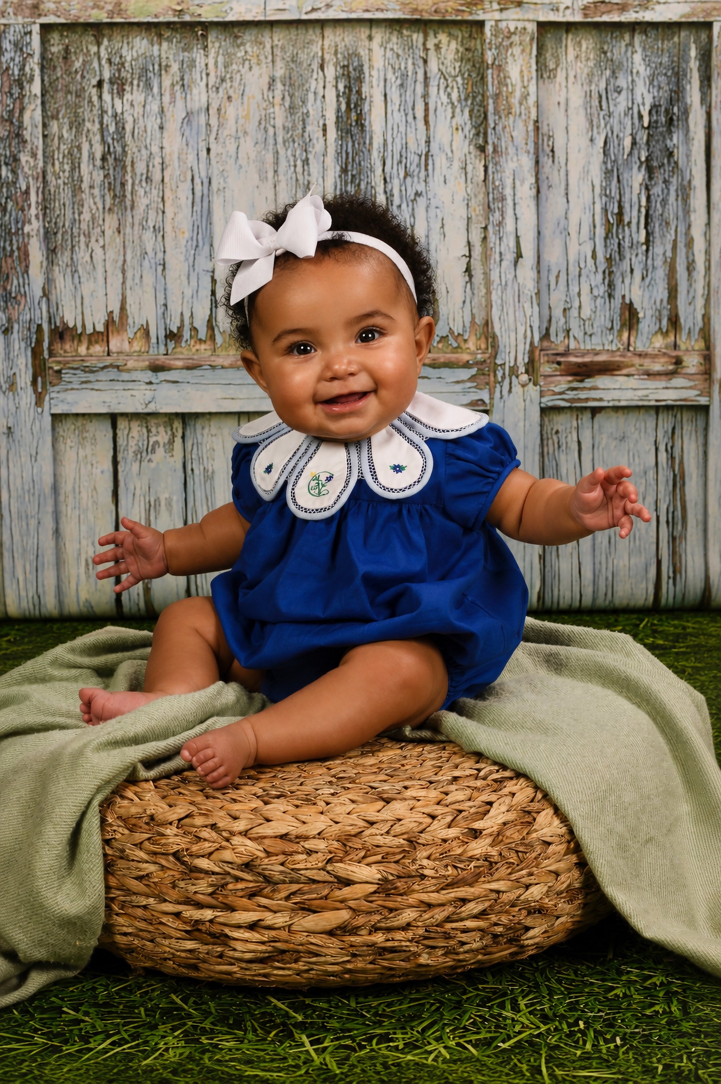 Adorable baby girl with curly hair, white bow headband, and blue dress, sitting on a wicker basket with a green cloth, smiling in front of a rustic, weathered wooden fence.