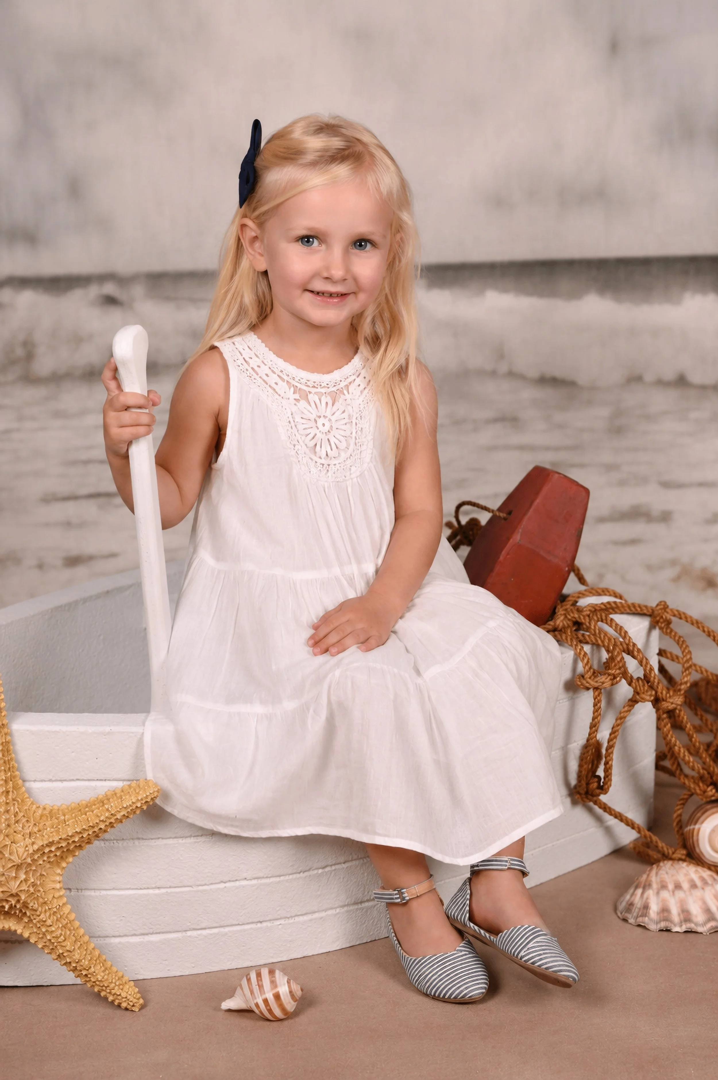 Young girl sitting on a boat, holding an oar, dressed in a white dress with lace details, wearing striped shoes, with seashells, a starfish, a red buoy, and knotted ropes around her, with a nautical background.