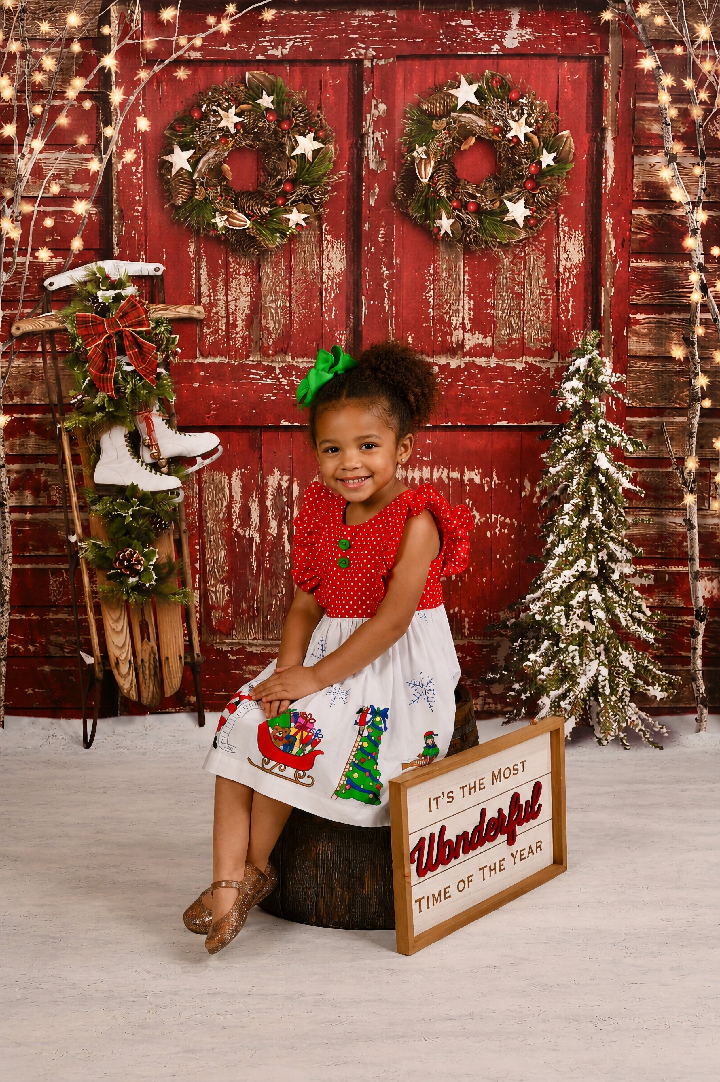 A young girl with curly hair in puffs, wearing a red and white Christmas dress with holiday-themed designs, is sitting on a barrel next to a wooden sign that reads 'It's the most wonderful time of the year.' She is smiling and has a green bow in her 