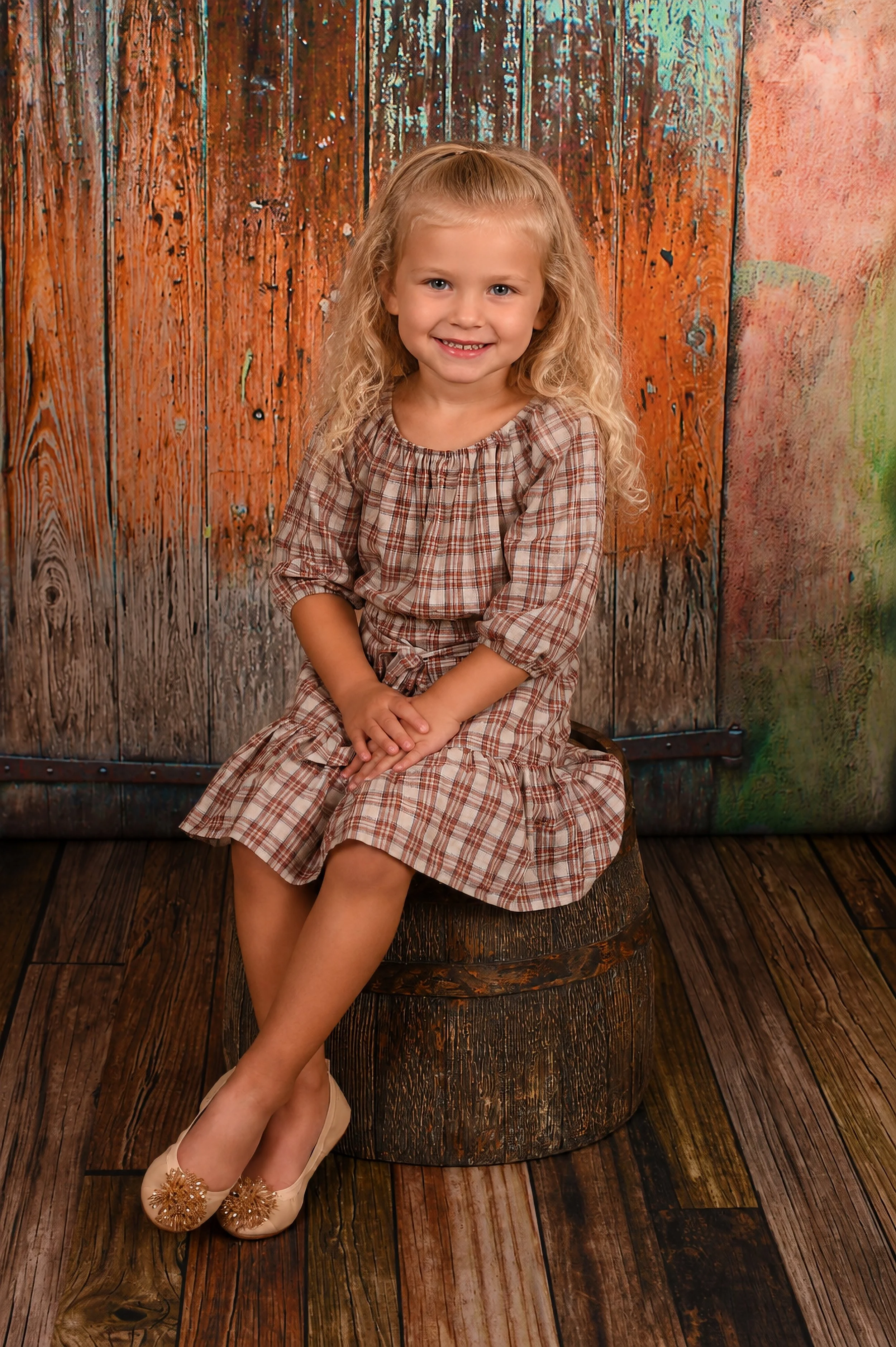 A young girl with curly blonde hair sitting on a wooden stool in front of a rustic wood and painted backdrop, smiling at the camera, wearing a brown plaid dress and cream-colored shoes with flower embellishments.