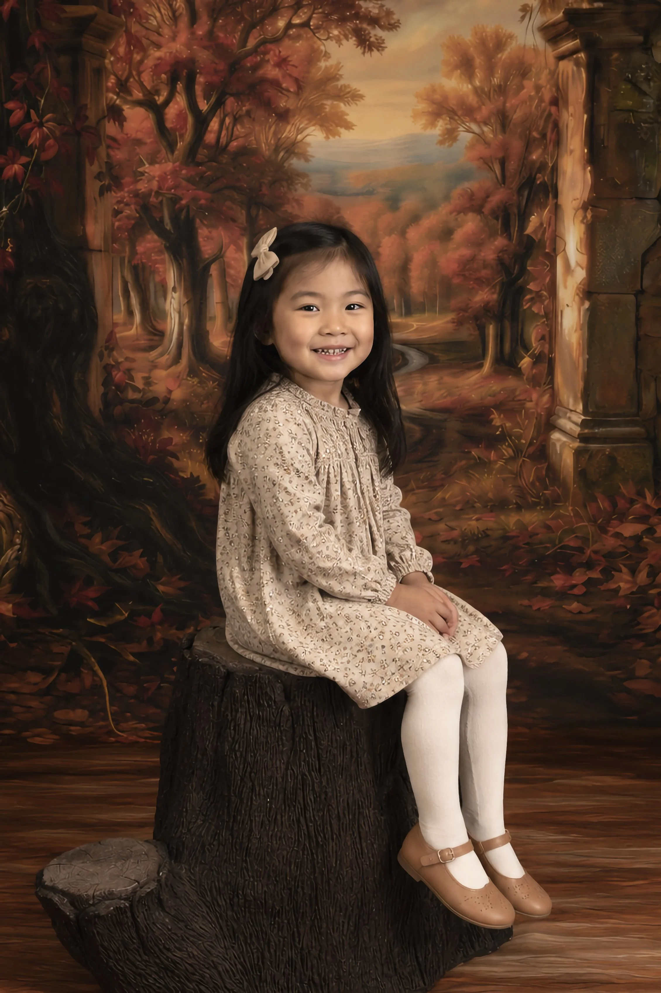 A young girl with long dark hair, wearing a beige lace dress, white tights, and tan shoes, sitting on a tree stump. She has a light-colored bow in her hair and is smiling. The background is a painted autumn landscape with trees and a winding path.
