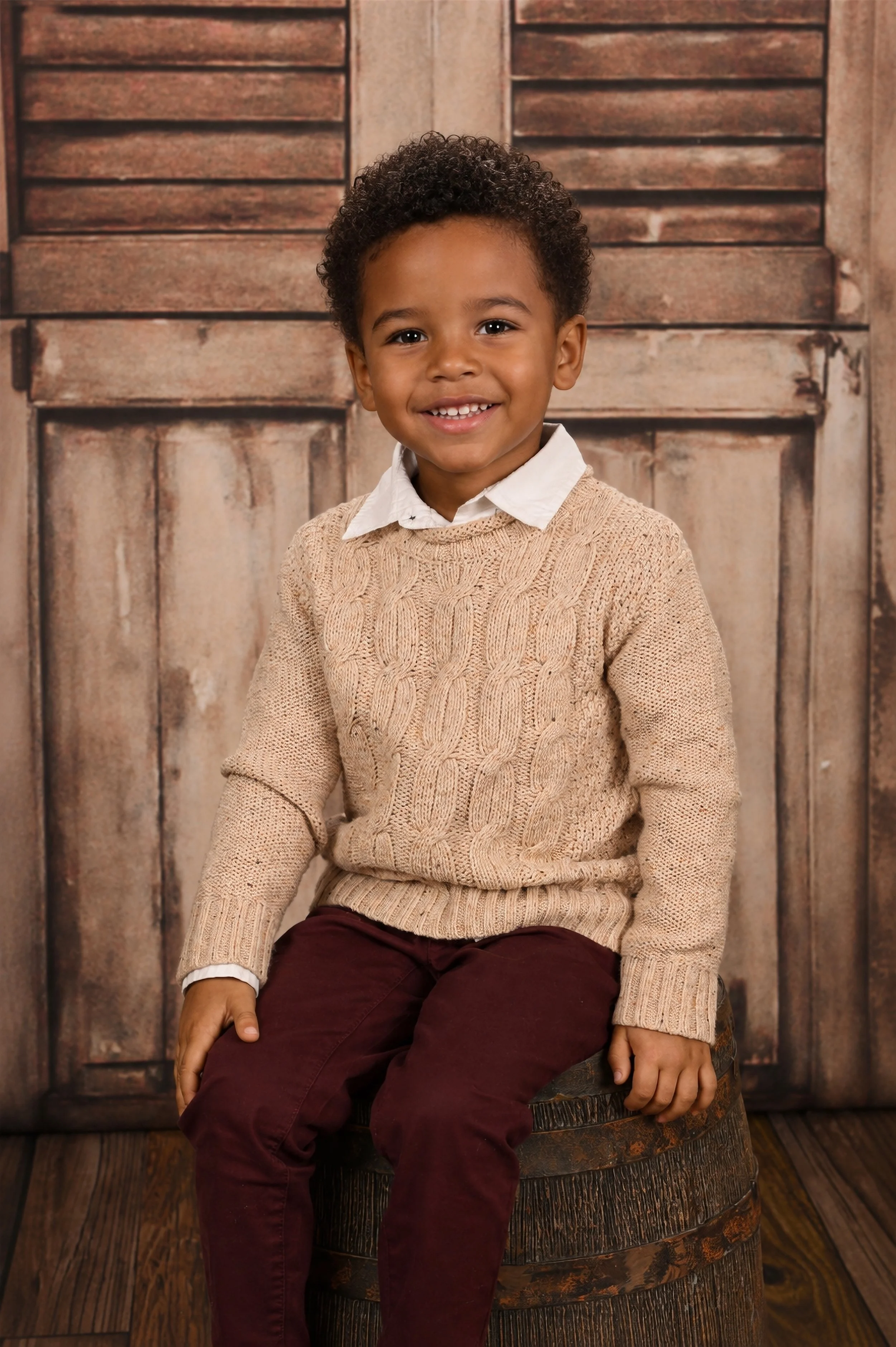 A young boy with curly hair, smiling, wearing a cream cable-knit sweater over a white collared shirt, burgundy pants, sitting on a wooden barrel in front of a rustic wooden background.