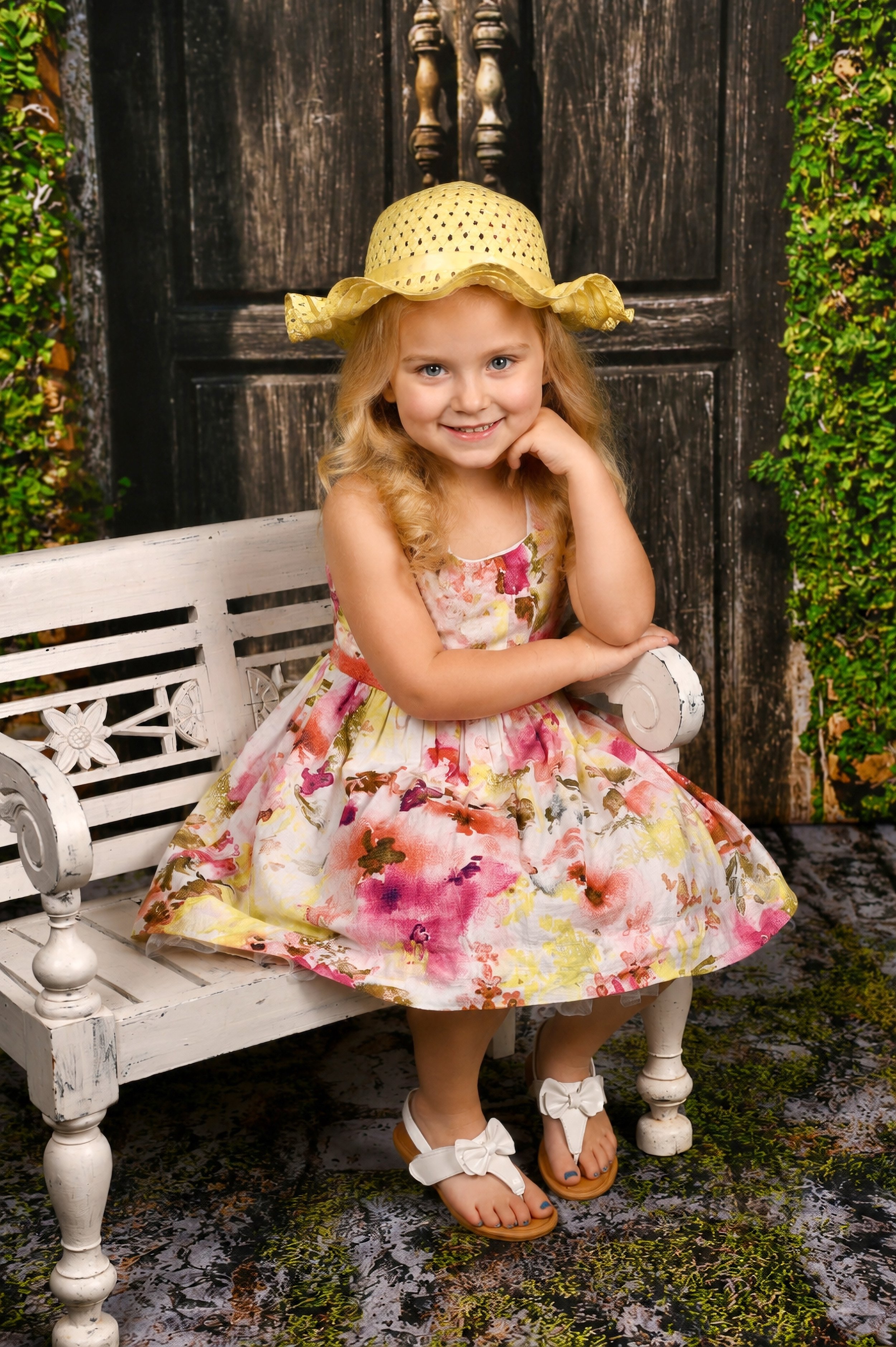A young girl with blonde hair, wearing a floral dress and white sandals with bows, sitting on a white bench outdoors. She is smiling, resting her chin on her hand, and wearing a yellow sun hat. Behind her is a black wooden door and greenery on the si