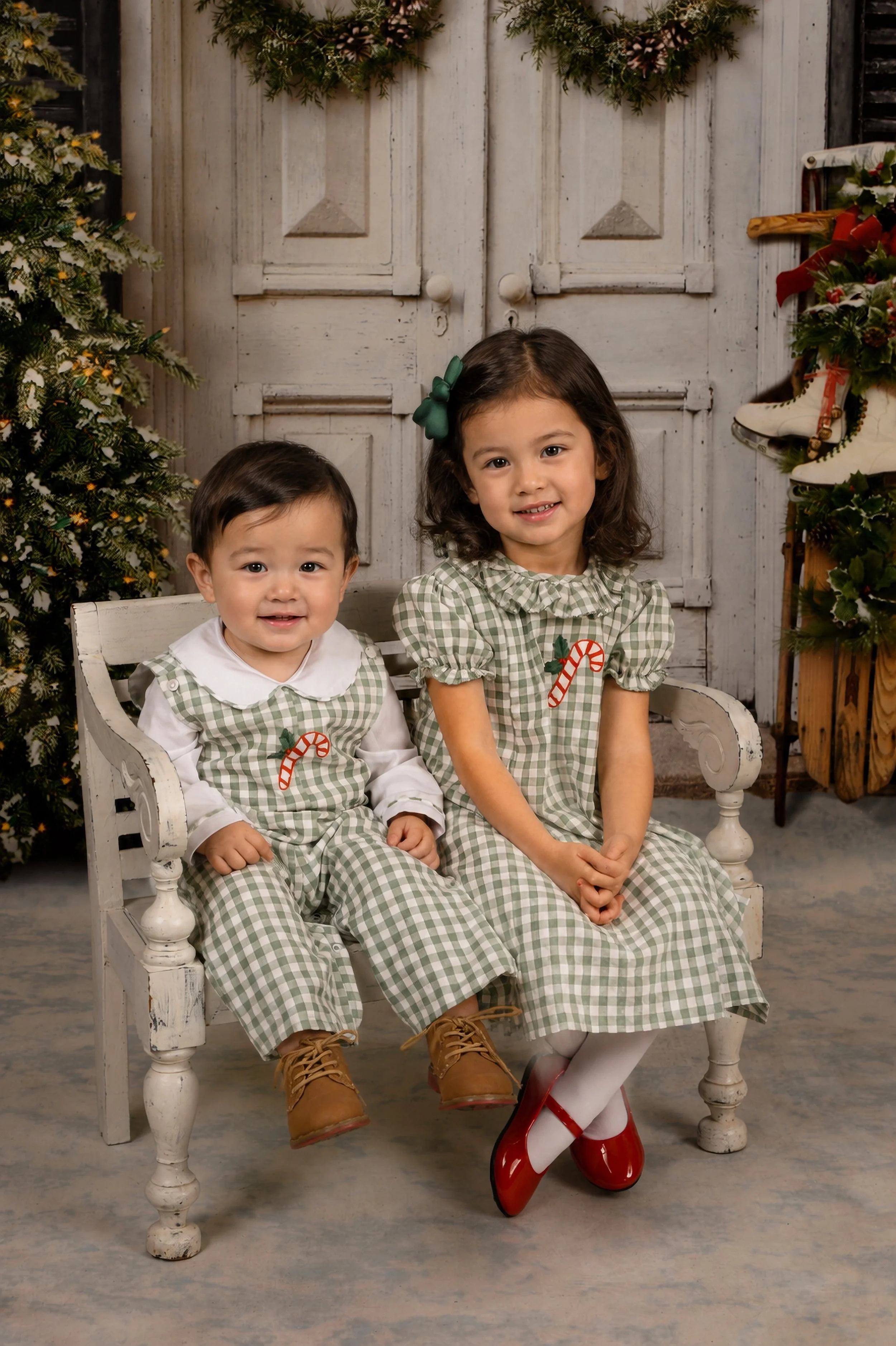 Two young children, a boy and a girl, sitting on a white wooden bench, dressed in matching green and white checkered Christmas outfits with candy cane embroidered on the front, in a holiday setting with decorated Christmas trees and Christmas-themed 