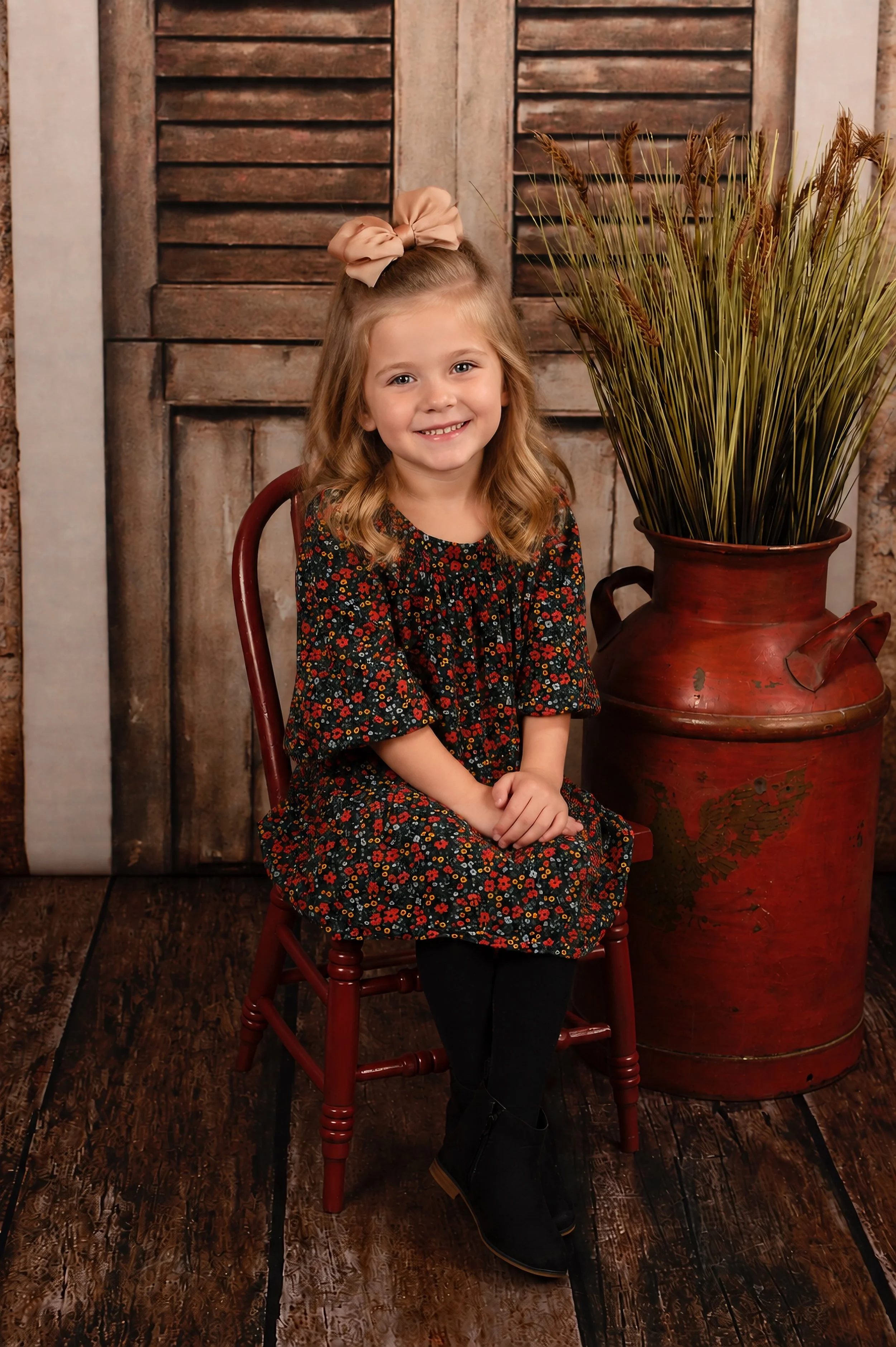 A young girl with blonde hair and a big beige bow, smiling, sitting on a red wooden chair, wearing a black floral dress with red, orange, and yellow flowers, black tights, and black boots, next to a large terracotta vase with dried wheat stalks, in f