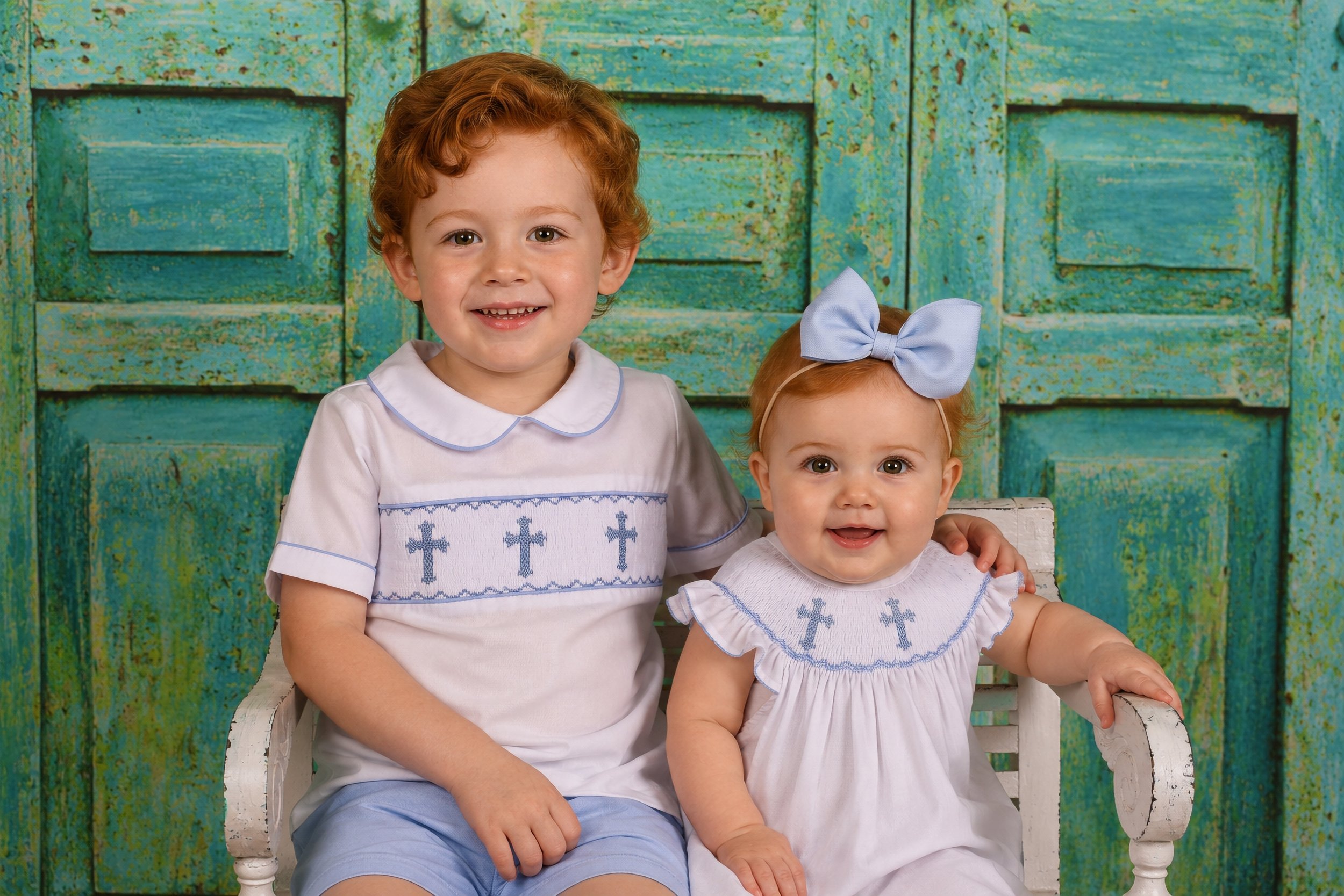 Two young girls sitting on a white bench in front of a weathered teal wooden background. The older girl has curly red hair, light skin, and is wearing a white dress with blue crosses. The younger girl has red hair, light skin, a large blue bow in her