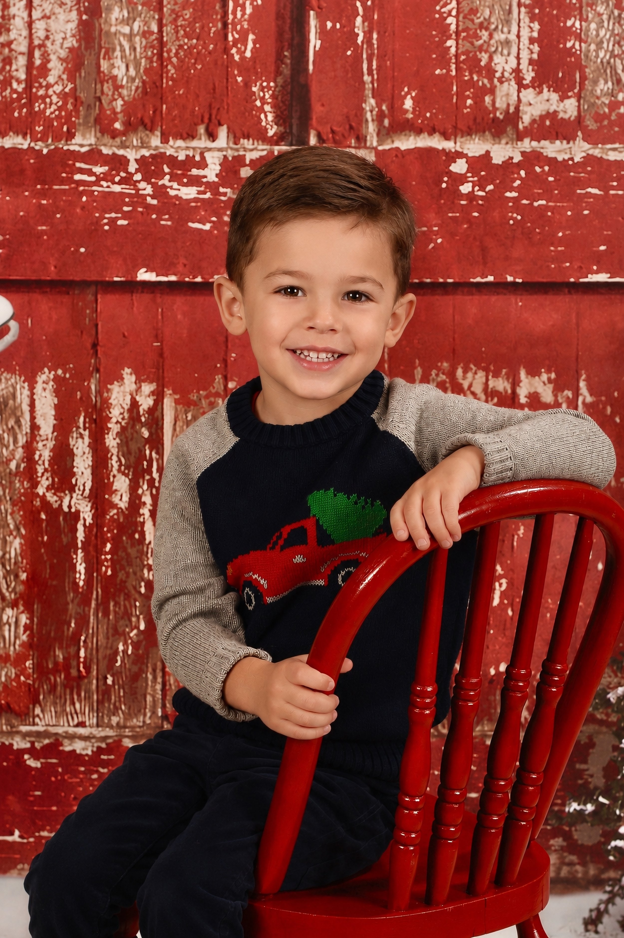 A young boy with brown hair and a big smile sitting on a red wooden chair in front of a distressed red wooden background, wearing a black and gray sweater with a Christmas tree design.