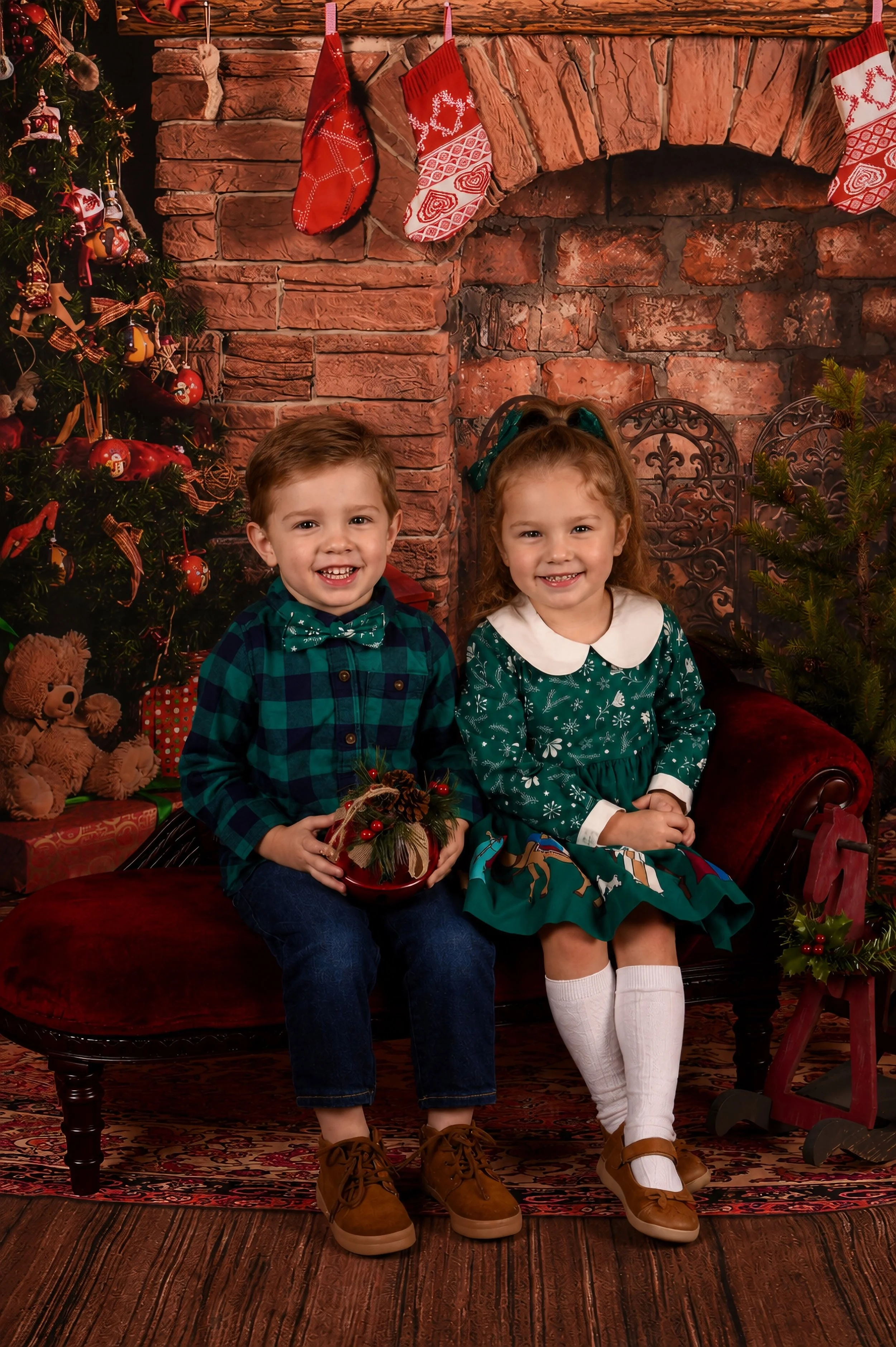 Two children, a boy and a girl, sitting on a red velvet bench in front of a brick fireplace with Christmas stockings hanging. The boy has short brown hair, is smiling, and wearing a green plaid shirt with a matching bow tie, dark jeans, and brown boo