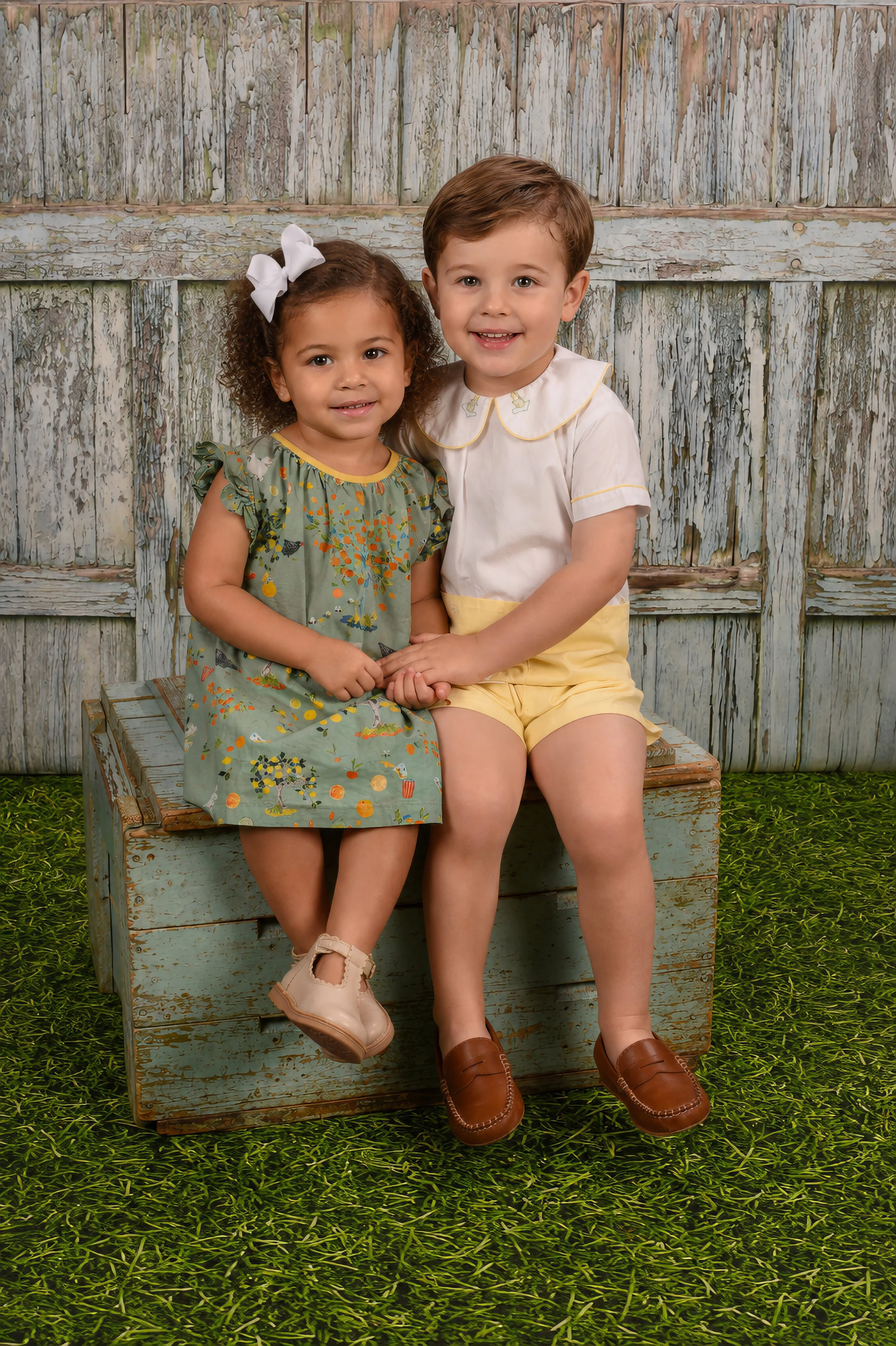Two young children, a girl and a boy, sitting on a rustic wooden bench, holding hands, with a weathered wooden fence behind them and green grass beneath.