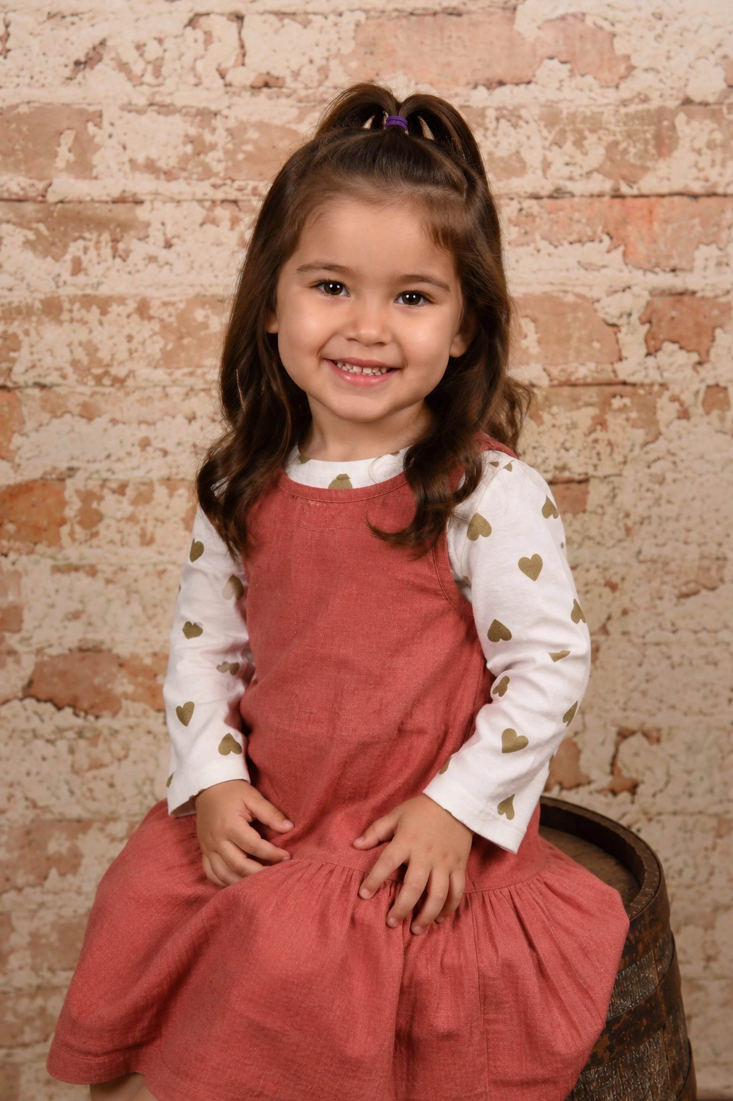 A young girl with brown hair in a high ponytail, sitting on a vintage wooden chair with a floral pattern, smiling at the camera. She is wearing a white long-sleeve shirt with gold heart prints and a red sleeveless dress. The background is a textured 