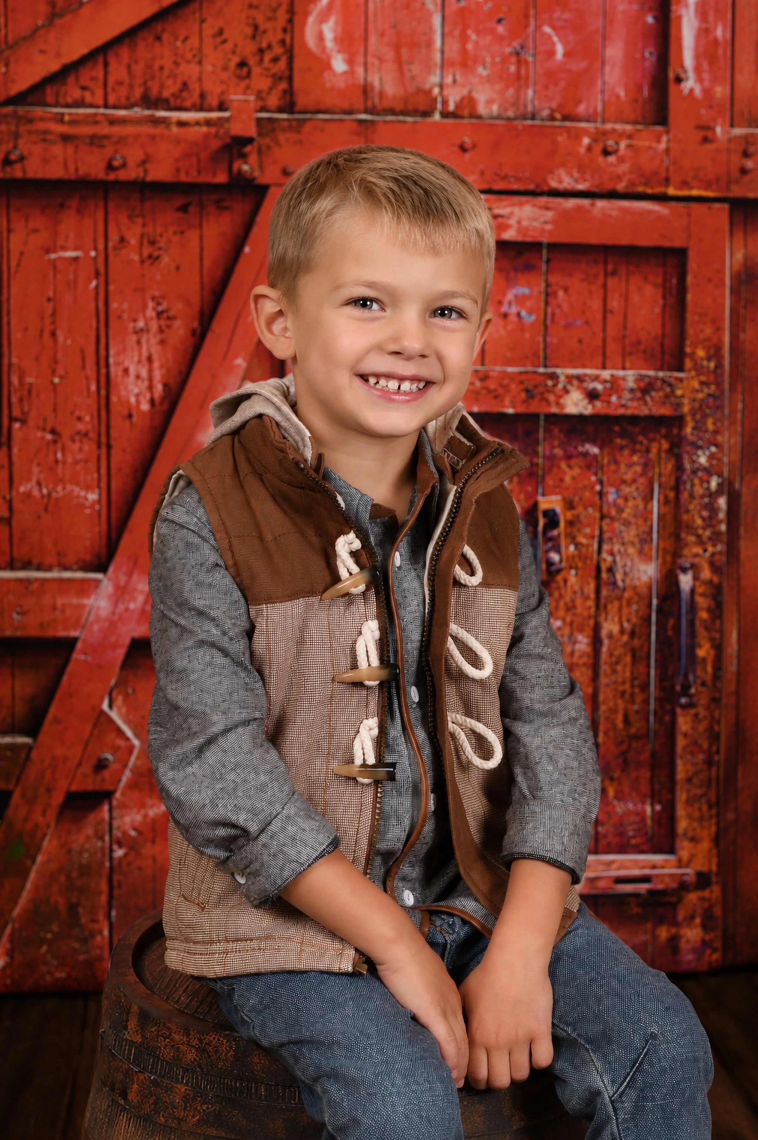 Young boy with blond hair smiling, sitting on a wooden barrel, wearing a gray shirt and brown vest, in front of a rustic red barn door.