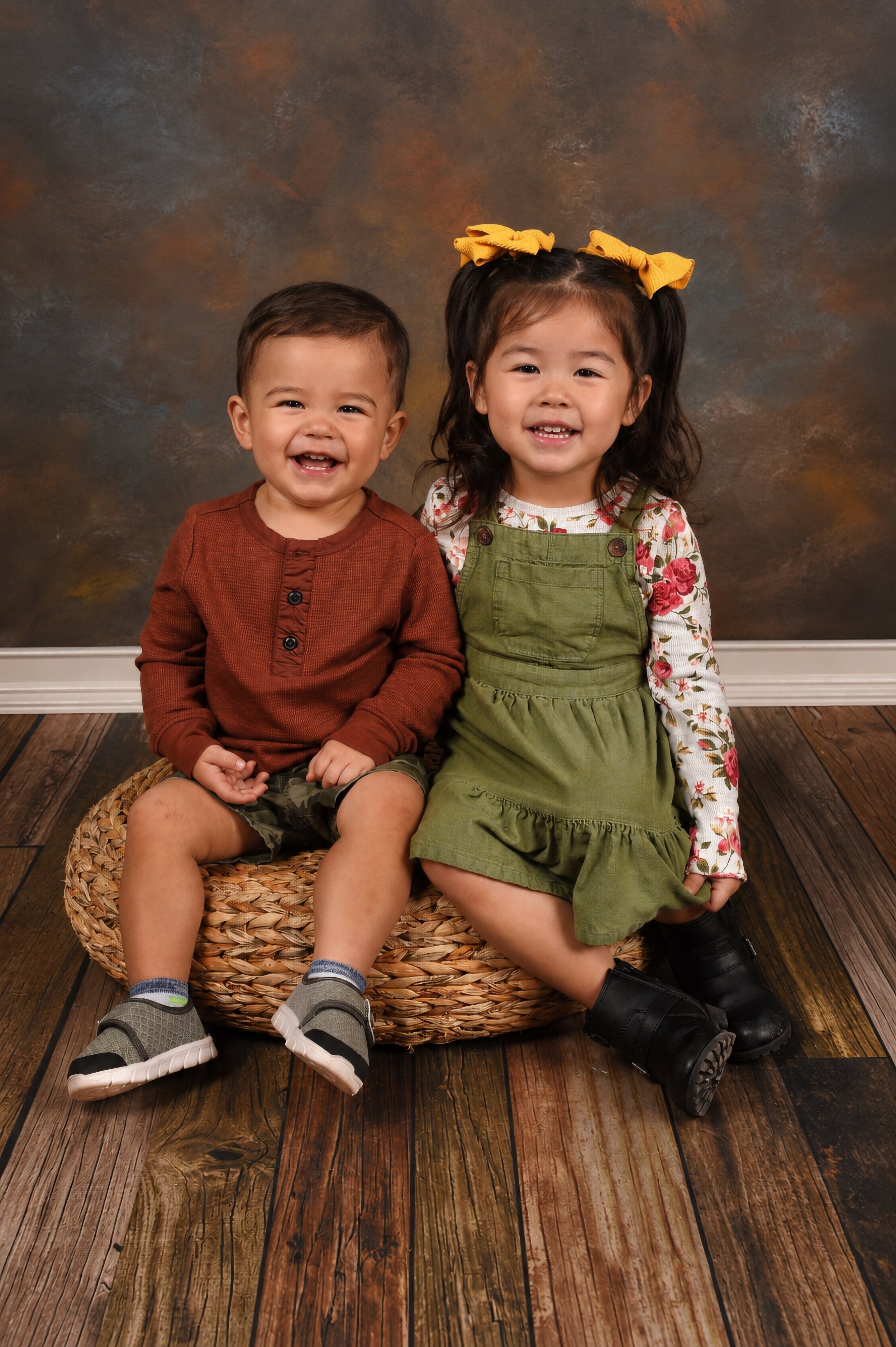 A young boy and girl sitting closely together on a woven basket, smiling against a brownish textured backdrop. The boy is wearing a rust-colored long-sleeve shirt, camo shorts, and gray sneakers with blue socks. The girl is dressed in a floral long-s