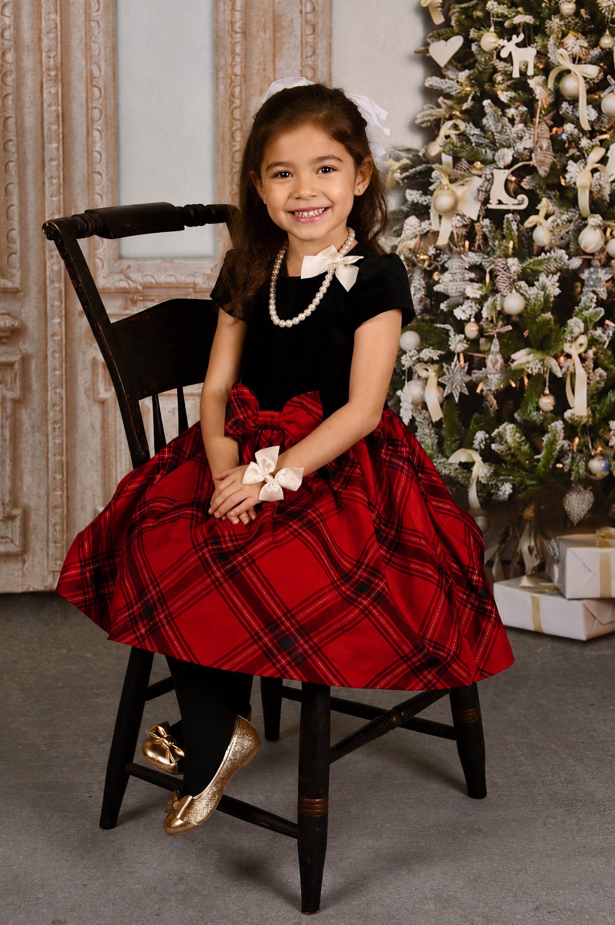 A young girl in a festive holiday outfit, wearing a black dress with a red plaid skirt, pearl necklace, and hair bow, sitting on a black wooden chair in front of a decorated Christmas tree with Christmas gifts underneath.