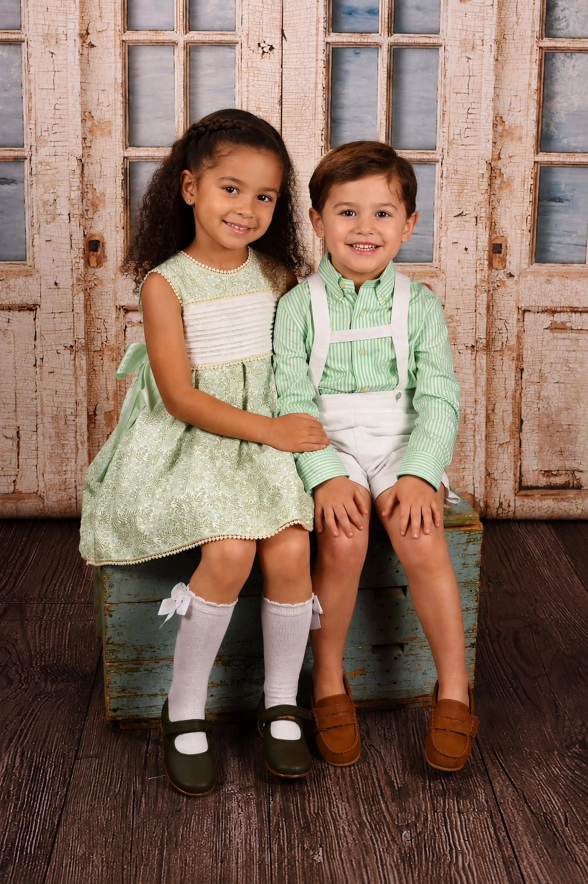 A young girl and boy sitting together on a vintage wooden box, smiling. The girl has curly hair and wears a green dress with white knee-high socks and black shoes. The boy has short brown hair and wears a green striped shirt with white suspenders, wh