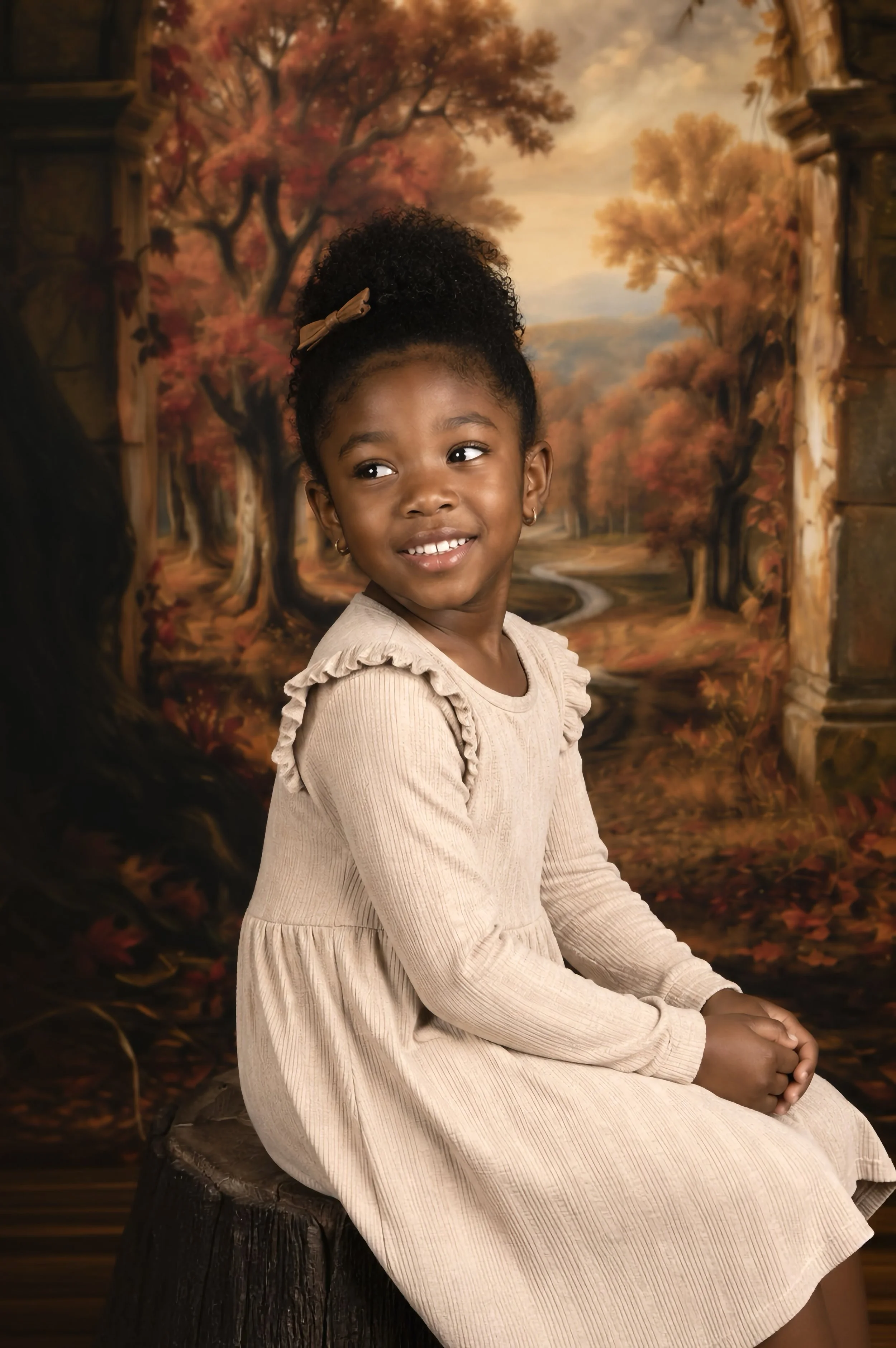Young girl with curly hair styled in a bun, wearing a beige long-sleeve dress with ruffled shoulders, sitting on a tree stump with a painted autumn scene background featuring trees with orange and red leaves and a winding path.