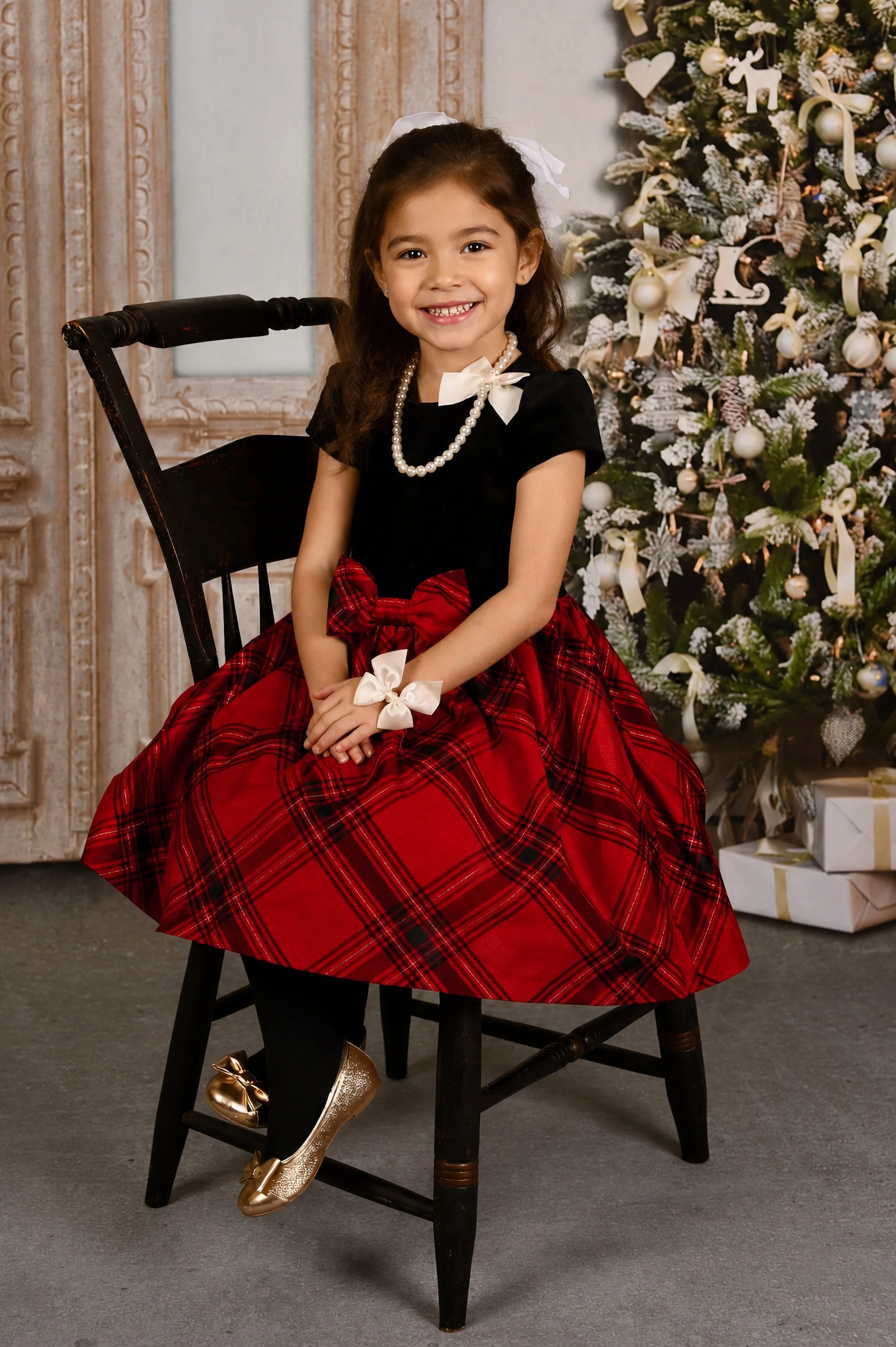 Young girl sitting on a black wooden chair, smiling in front of a decorated Christmas tree with presents underneath, wearing a red plaid dress, black shoes with gold accents, a pearl necklace, and a white hair bow.