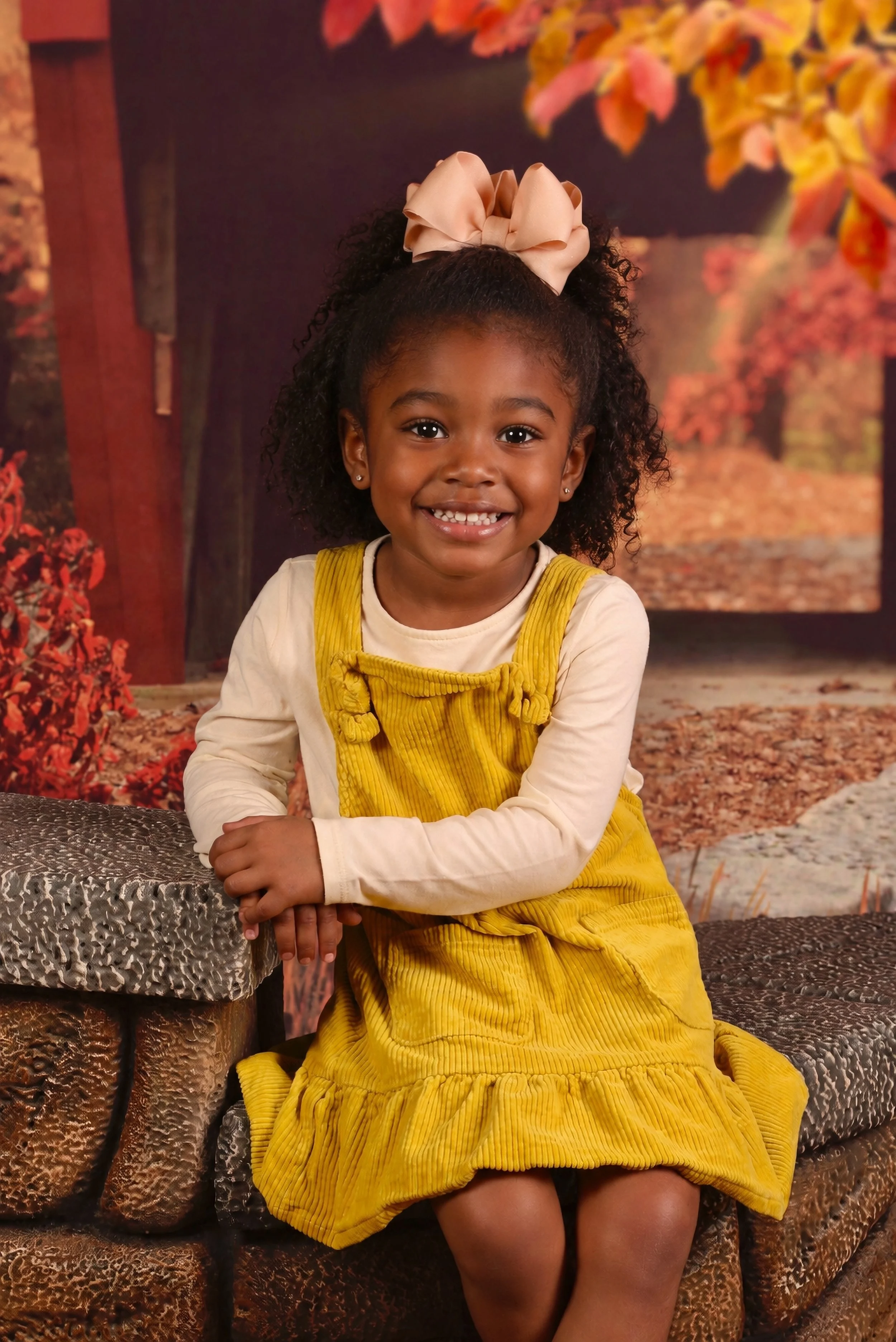 A young girl with dark curly hair, wearing a large beige bow in her hair, a cream long-sleeve shirt, and a yellow corduroy dress, sitting on a patterned couch outdoors in autumn setting with trees and falling leaves in the background, smiling at the 