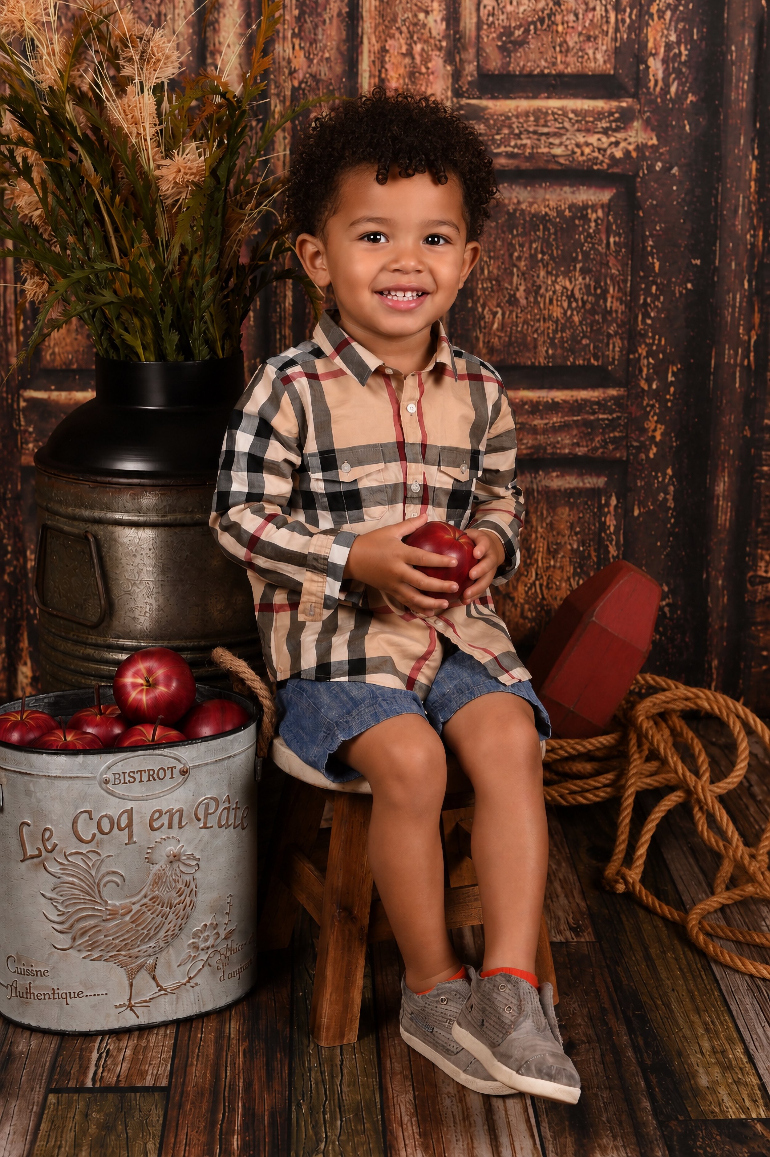 A young boy with curly hair, wearing a plaid shirt and denim shorts, sitting on a wooden stool while holding a red apple. He's smiling at the camera with a rustic background, which includes a dark wooden door, a potted plant, an antique metal milk ca