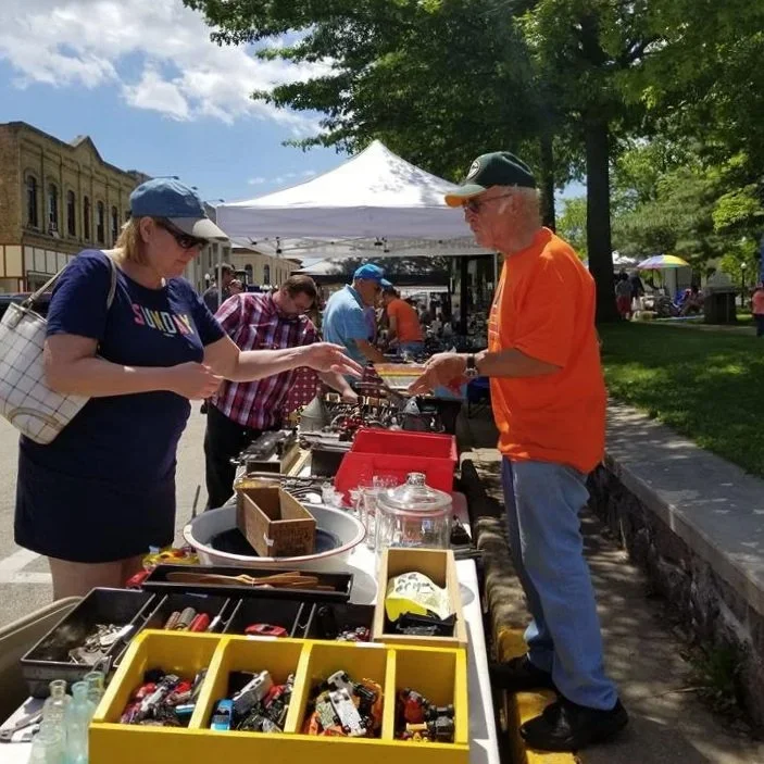People browsing vintage and toy collectible items at an outdoor market with tents and trees under a sunny sky.