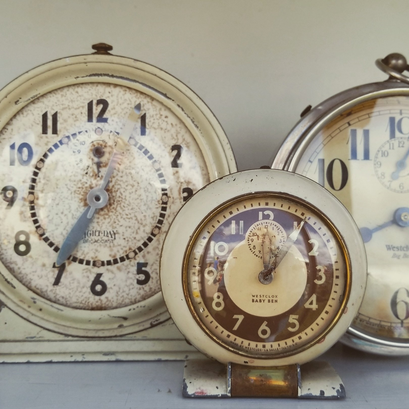 Close-up image of vintage clocks, including a large clock with black numbers and a rusty frame, a smaller clock labeled 'Westclox Baby Ben', and part of another clock in the background.