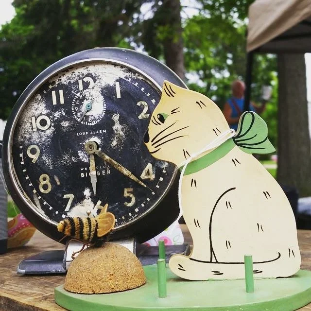 A vintage clock, vintage cat decor wearing a green collar beside it, a small bee figure are possible items found at our outdoor market on Sundays.