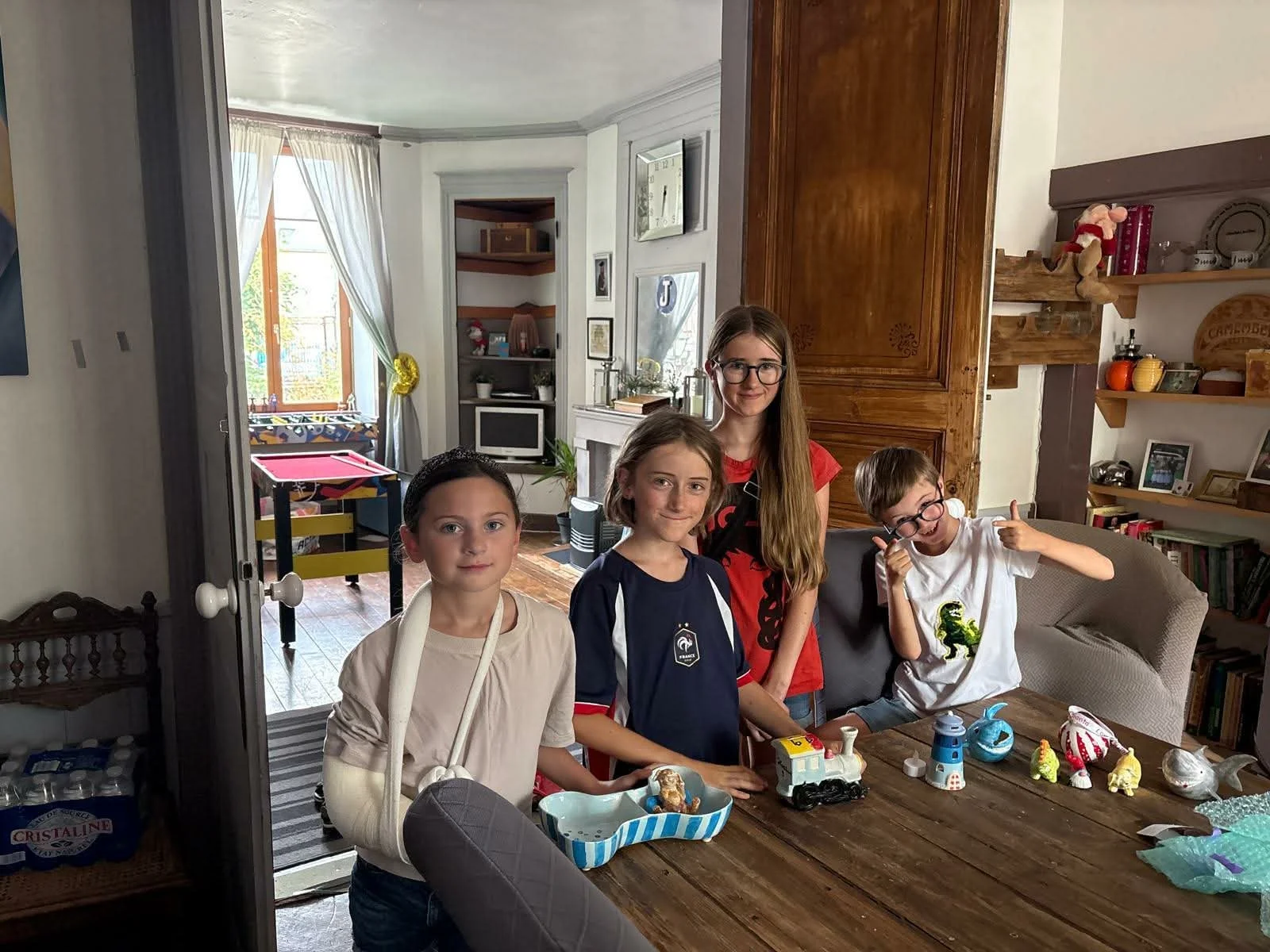 Four children standing around a table with toys and treats inside a house, with a room and large window in the background.