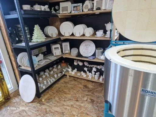 Display of white ceramic plates, bowls, and decorative items on black shelves in a store. There is a large ice cream maker on the right side.