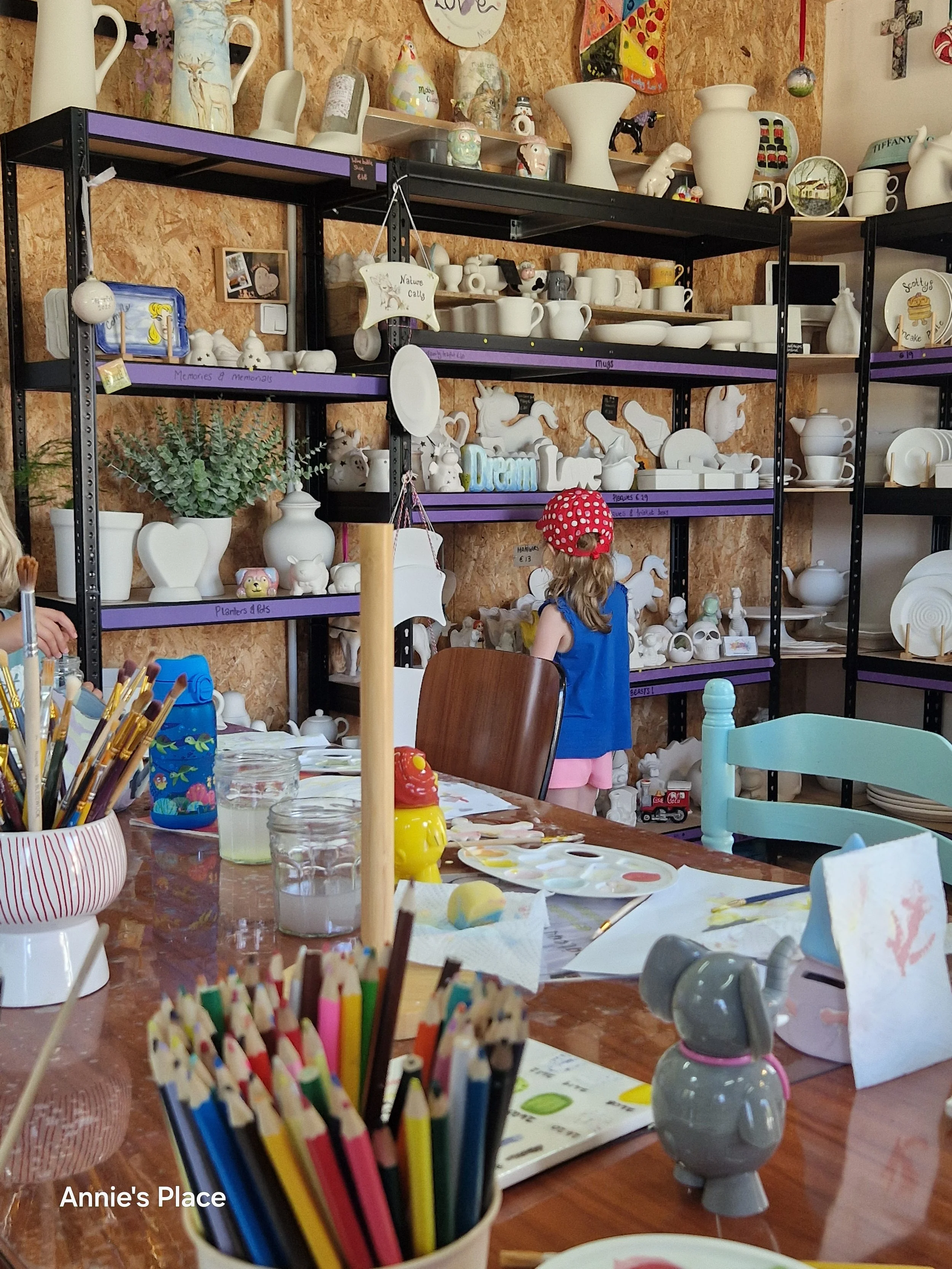 A craft or art workspace with shelves stocked with various ceramic and porcelain figurines, mugs, vases, and decorative items. A young girl wearing a red polka-dot hat and a blue shirt is seen from behind, working at the table which has art supplies such as colored pencils, paint, and paper. The space has a rustic look with wooden walls and organized shelves.