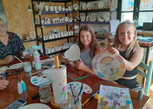 Two young girls smiling and holding painted ceramic items, surrounded by painting supplies in a ceramics painting studio.