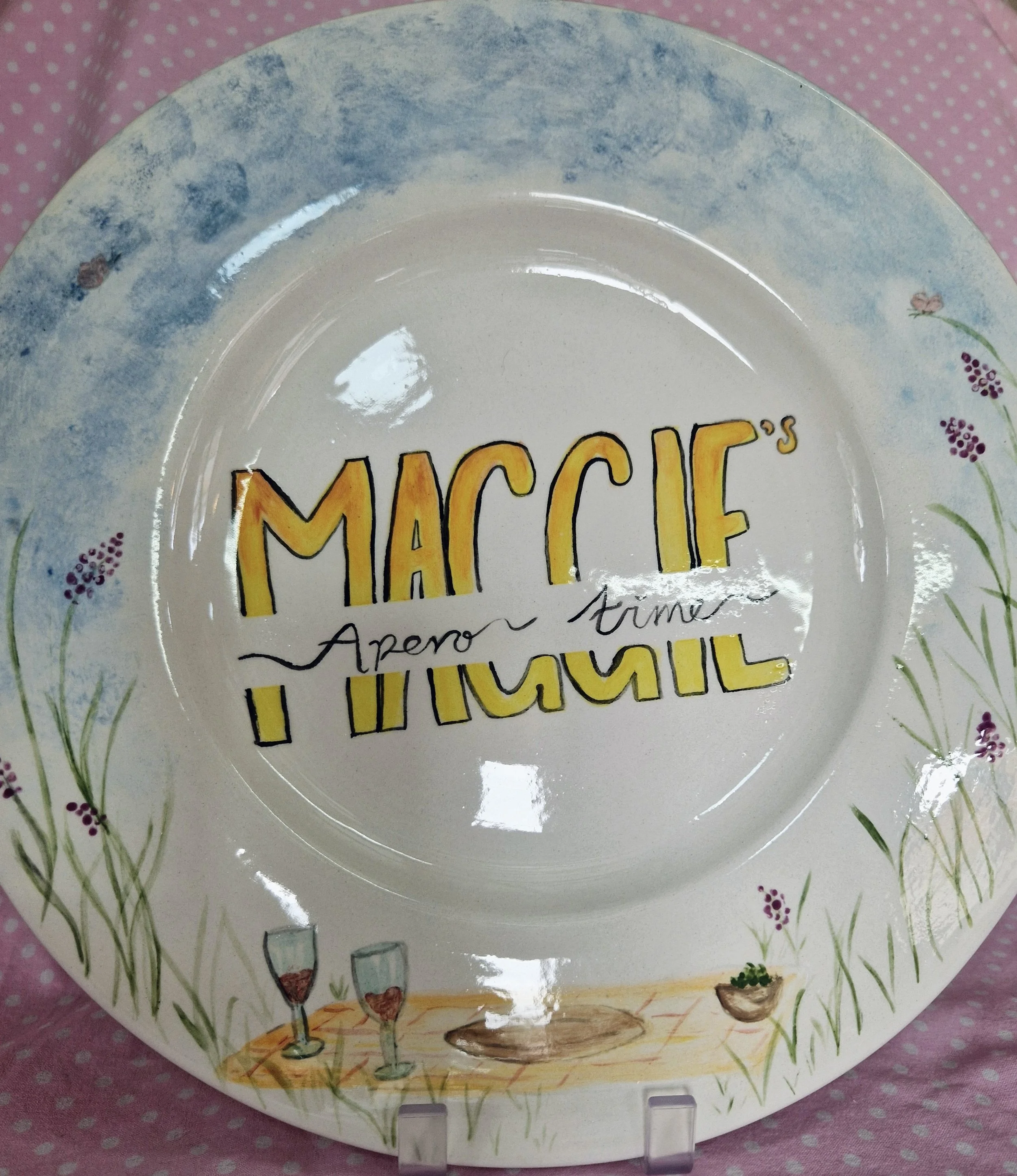 Decorative ceramic plate with watercolor illustrations of wine glasses, plants, and pink flowers, resting on a pink polka-dotted surface.